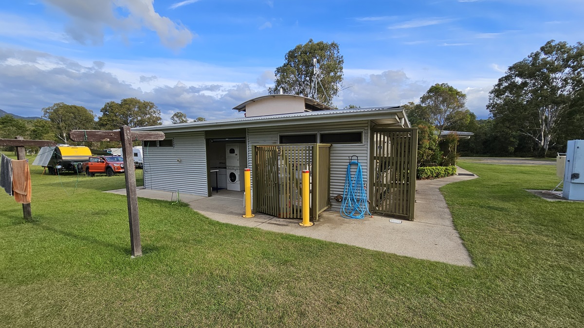 A small, single-storey building with a metal exterior and a flat roof is in the centre. It has an open entrance revealing a washing machine inside. To the right, a blue hose is coiled on the wall. Yellow bollards are in front of a wooden screen. On the left, a clothesline with towels is visible. In the background, there are trees and a few parked vehicles, including a yellow trailer and an orange car. The ground is covered with grass. A small, single-storey building with a metal exterior and a flat roof is in the centre. It has an open entrance revealing a washing machine inside. To the right, a blue hose is coiled on the wall. Yellow bollards are in front of a wooden screen. On the left, a clothesline with towels is visible. In the background, there are trees and a few parked vehicles, including a yellow trailer and an orange car. The ground is covered with grass.