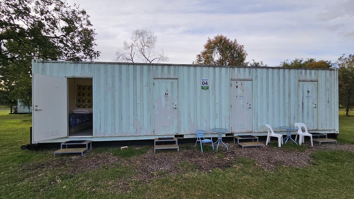 A long, light blue shipping container with four doors, each accessed by small metal steps. The leftmost door is open, revealing a glimpse of the interior. In front of the container, there are two white plastic chairs and a blue metal chair, along with a small table. The container is set on a grassy area with some trees in the background. The ground in front of the container is covered with patches of gravel. A long, light blue shipping container with four doors, each accessed by small metal steps. The leftmost door is open, revealing a glimpse of the interior. In front of the container, there are two white plastic chairs and a blue metal chair, along with a small table. The container is set on a grassy area with some trees in the background. The ground in front of the container is covered with patches of gravel.