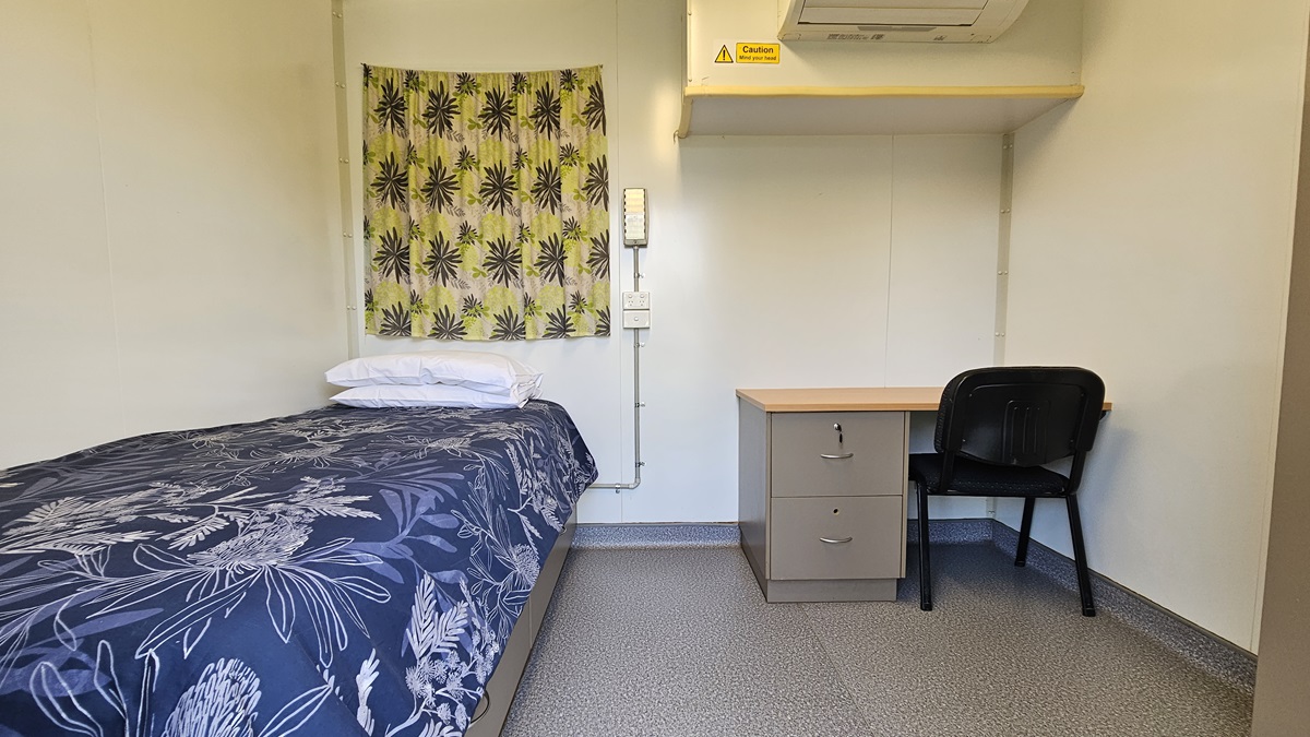 A small room with a single bed on the left, covered with a dark blue floral-patterned duvet and white pillows. Above the bed is a yellow curtain with a black starburst design. On the right, there's a wooden desk with a black chair. Above the desk is an air conditioning unit with a caution sign. The floor is grey and speckled, and the walls are plain white. A small room with a single bed on the left, covered with a dark blue floral-patterned duvet and white pillows. Above the bed is a yellow curtain with a black starburst design. On the right, there's a wooden desk with a black chair. Above the desk is an air conditioning unit with a caution sign. The floor is grey and speckled, and the walls are plain white.