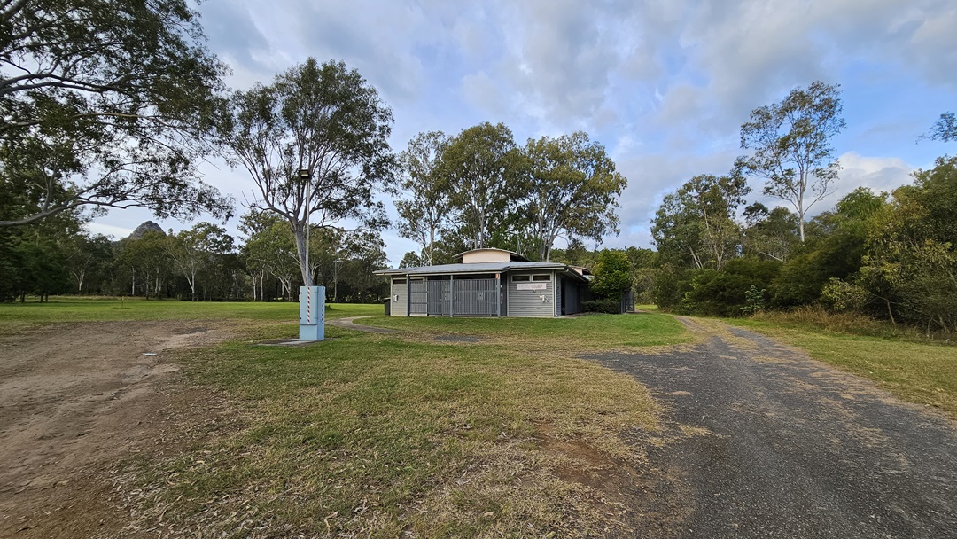 A modern, single-storey building with a flat roof and grey slatted exterior. It is surrounded by a grassy area with a few small bushes in front. On the left and right sides, there are rubbish bins near the building. In the background, there are tall trees with green foliage. The sky is overcast. A modern, single-storey building with a flat roof and grey slatted exterior. It is surrounded by a grassy area with a few small bushes in front. On the left and right sides, there are rubbish bins near the building. In the background, there are tall trees with green foliage. The sky is overcast.