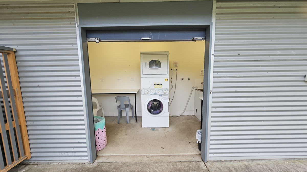 A small laundry room with corrugated metal walls. In the centre, there's a white stacked washer and dryer. To the left, a plastic chair and a laundry basket with a polka dot design. On the right, a countertop with a sink and some plumbing fixtures. The floor is concrete, and the ceiling is not visible. A small laundry room with corrugated metal walls. In the centre, there's a white stacked washer and dryer. To the left, a plastic chair and a laundry basket with a polka dot design. On the right, a countertop with a sink and some plumbing fixtures. The floor is concrete, and the ceiling is not visible.