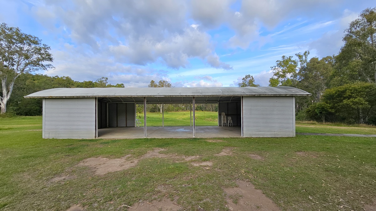 A simple, open-sided structure with a corrugated metal roof and walls stands on a grassy area. The building has three open sections supported by metal poles, with the middle section providing a view of the green landscape beyond. Inside, there are a few stools visible on the right. Trees and bushes surround the area, creating a natural backdrop. The ground is a mix of grass and patches of bare earth. A simple, open-sided structure with a corrugated metal roof and walls stands on a grassy area. The building has three open sections supported by metal poles, with the middle section providing a view of the green landscape beyond. Inside, there are a few stools visible on the right. Trees and bushes surround the area, creating a natural backdrop. The ground is a mix of grass and patches of bare earth.