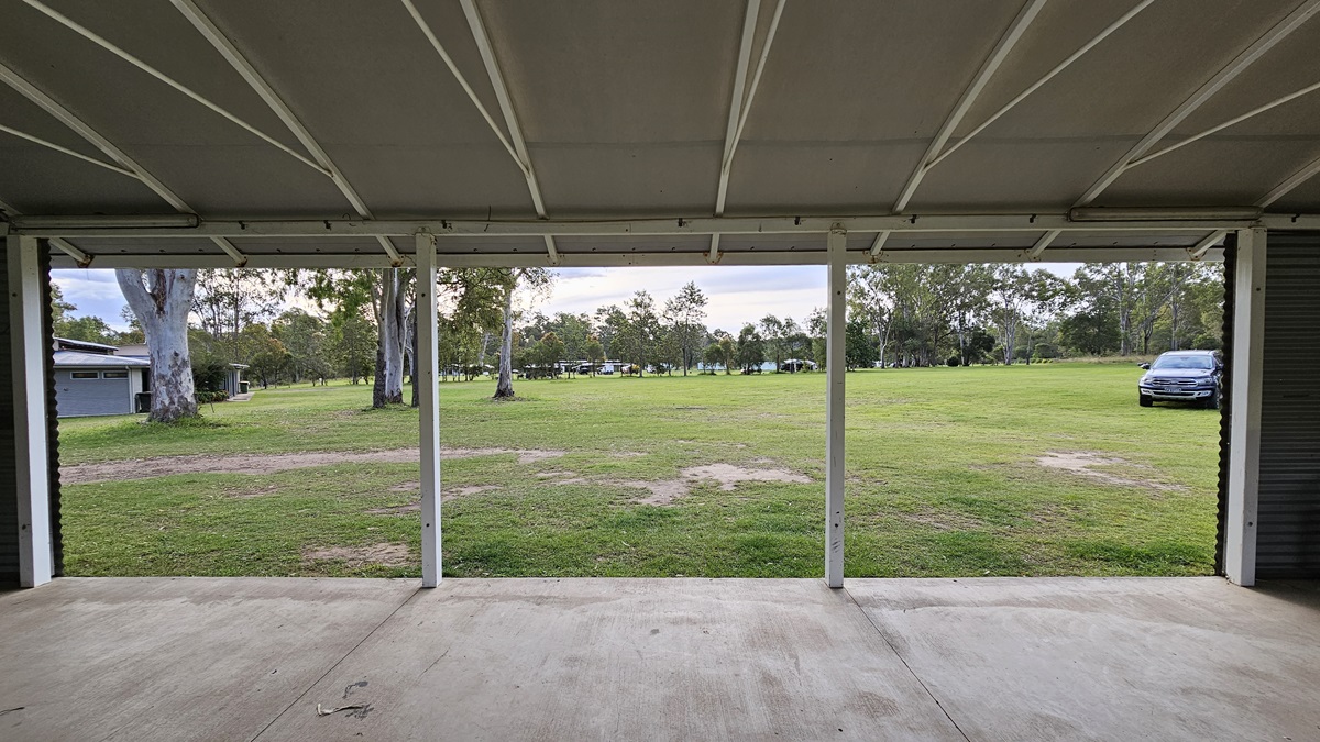 A view from inside a covered area with a concrete floor, looking out onto a grassy field. The ceiling is supported by white beams. In the background, there are several trees scattered across the field. To the right, a parked car is visible on the grass. On the left, a building is partially visible behind the trees. A view from inside a covered area with a concrete floor, looking out onto a grassy field. The ceiling is supported by white beams. In the background, there are several trees scattered across the field. To the right, a parked car is visible on the grass. On the left, a building is partially visible behind the trees.