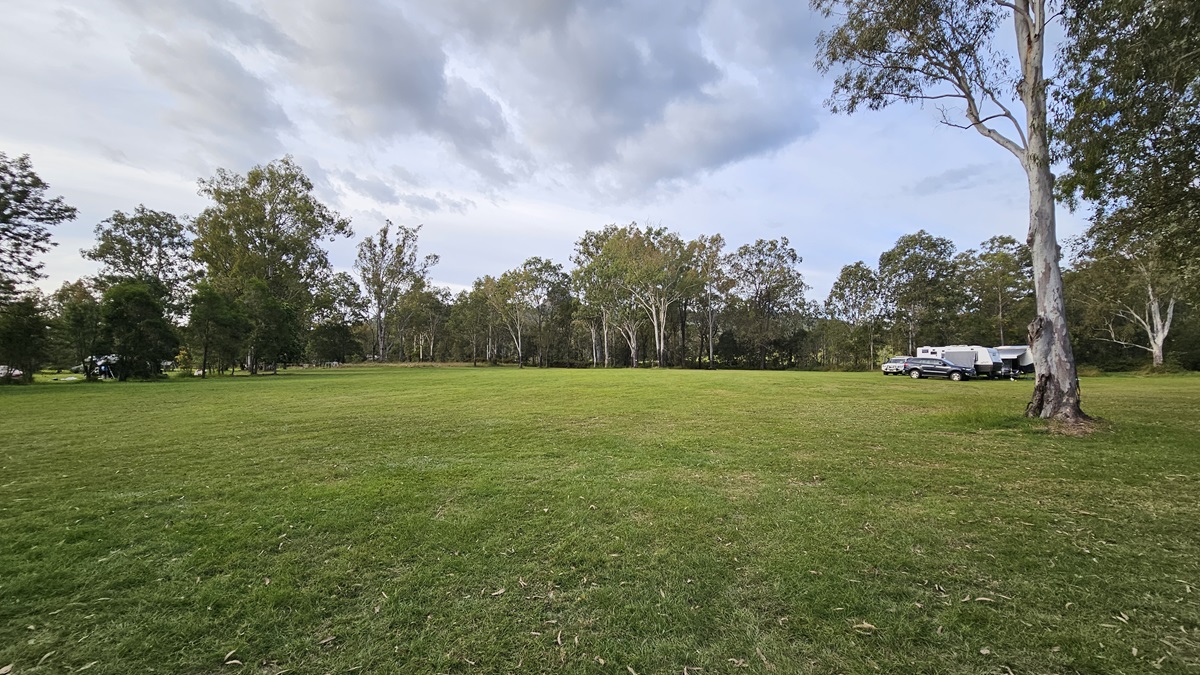 A grassy field stretches across the foreground, with a line of trees in the background. On the right, there are several parked caravans and a car near a large tree. The sky is overcast with grey clouds. A grassy field stretches across the foreground, with a line of trees in the background. On the right, there are several parked caravans and a car near a large tree. The sky is overcast with grey clouds.