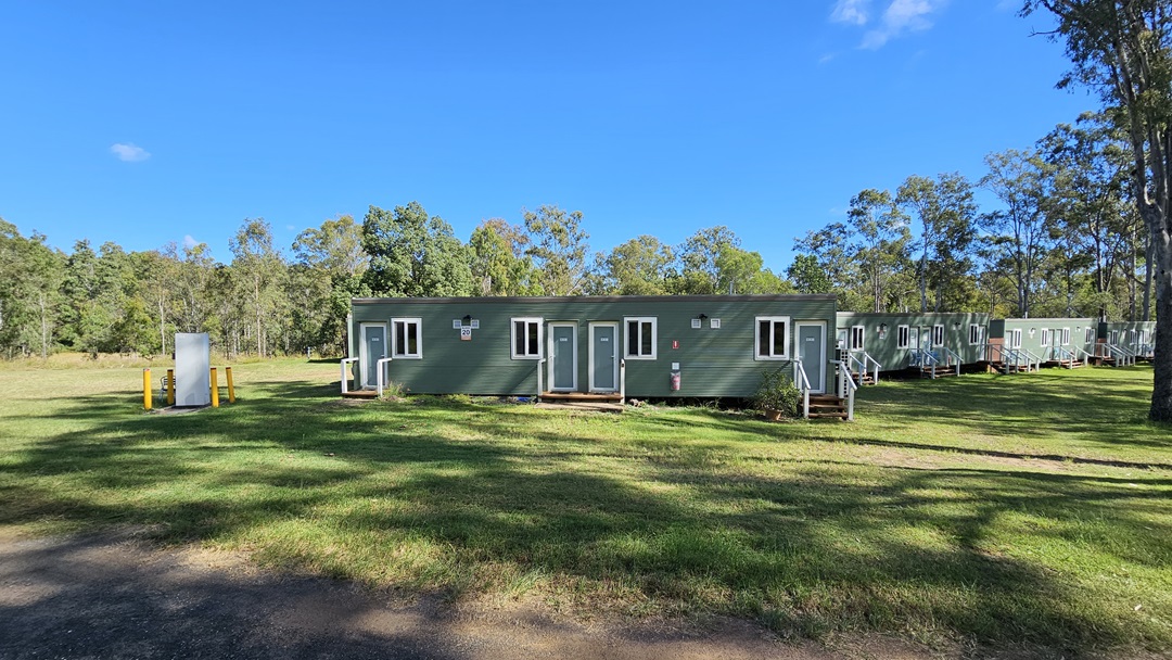 A dirt path runs through a grassy area, leading towards a row of long, green portable buildings in the background. Tall trees with slender trunks are scattered throughout the scene, casting shadows on the ground. The sky is clear and blue, and the area is surrounded by more trees, creating a wooded backdrop. A dirt path runs through a grassy area, leading towards a row of long, green portable buildings in the background. Tall trees with slender trunks are scattered throughout the scene, casting shadows on the ground. The sky is clear and blue, and the area is surrounded by more trees, creating a wooded backdrop.