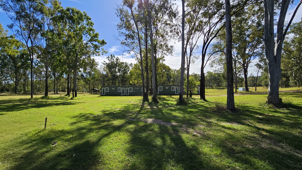 A grassy field with tall trees scattered throughout. In the middle, a long, single-storey building with multiple windows is visible. The shadows of the trees stretch across the grass, indicating sunlight from above. The background is filled with more trees, creating a wooded area. The scene is peaceful and open, with a clear view of the landscape. A grassy field with tall trees scattered throughout. In the middle, a long, single-storey building with multiple windows is visible. The shadows of the trees stretch across the grass, indicating sunlight from above. The background is filled with more trees, creating a wooded area. The scene is peaceful and open, with a clear view of the landscape.