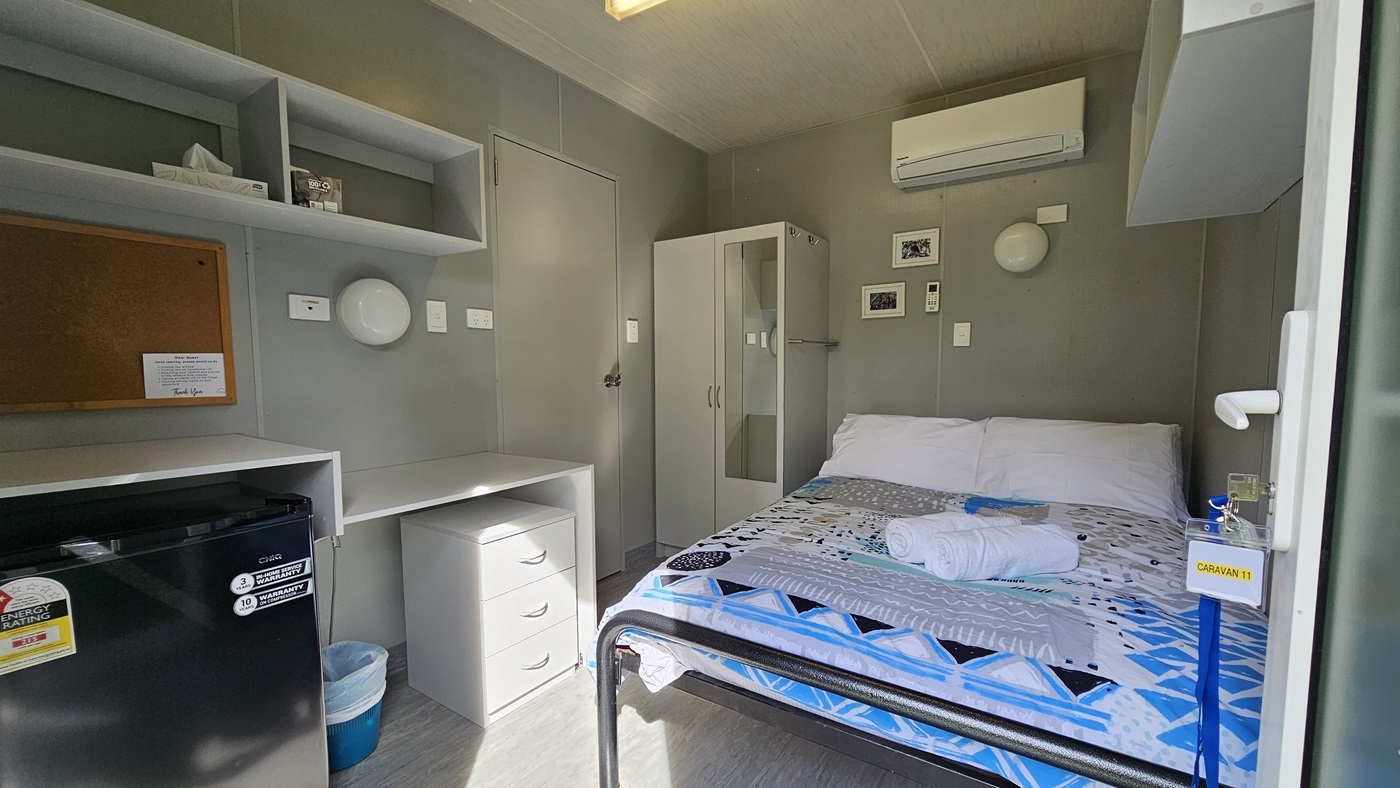 A compact room with a bed on the right, covered with a geometric-patterned bedspread and a towel on top. Above the bed is an air conditioning unit. On the left, there are shelves with a tissue box and a corkboard. Below the shelves is a small fridge and a set of drawers. A wardrobe is next to the bed. The walls are grey, and the floor is light-coloured. The ceiling has a fluorescent light fixture. A compact room with a bed on the right, covered with a geometric-patterned bedspread and a towel on top. Above the bed is an air conditioning unit. On the left, there are shelves with a tissue box and a corkboard. Below the shelves is a small fridge and a set of drawers. A wardrobe is next to the bed. The walls are grey, and the floor is light-coloured. The ceiling has a fluorescent light fixture.