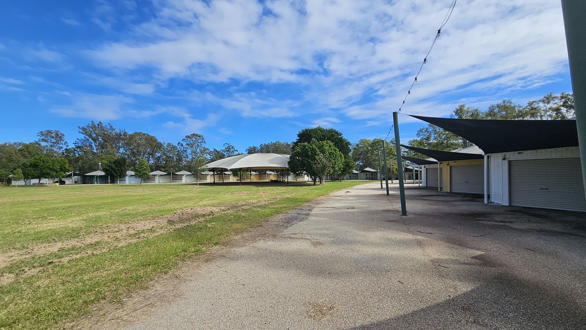 A grassy field on the left with a row of small, round huts in the distance. In the centre, a larger building with a grey roof is partially obscured by trees. On the right, a paved path runs alongside a series of closed stalls with dark shade sails overhead. The background features a line of trees under a partly cloudy sky. A grassy field on the left with a row of small, round huts in the distance. In the centre, a larger building with a grey roof is partially obscured by trees. On the right, a paved path runs alongside a series of closed stalls with dark shade sails overhead. The background features a line of trees under a partly cloudy sky.
