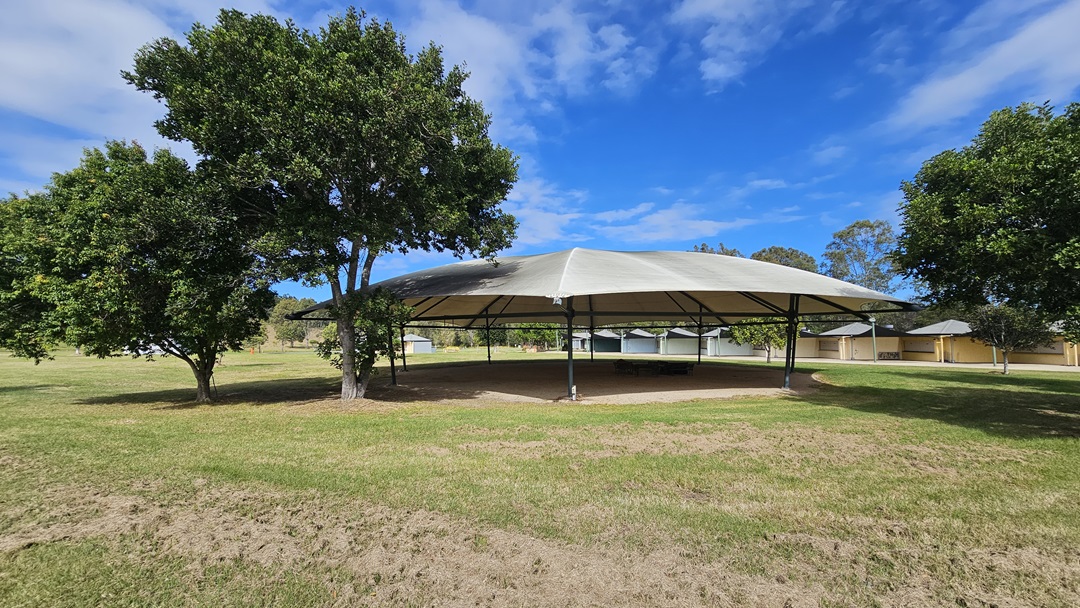 A large, open-sided pavilion with a white, tent-like roof is situated in the centre of a grassy area. On the left and right, there are several green trees providing shade. In the background, there are more pavilions and a few scattered trees. The ground is covered with grass, and the sky is partly cloudy. A large, open-sided pavilion with a white, tent-like roof is situated in the centre of a grassy area. On the left and right, there are several green trees providing shade. In the background, there are more pavilions and a few scattered trees. The ground is covered with grass, and the sky is partly cloudy.