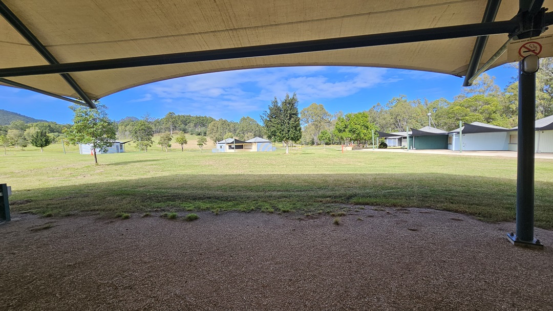A grassy field stretches across the middle of the image, with several small buildings scattered in the distance. Trees are visible in the background, providing a natural border. In the foreground, there is a gravel area under a large, curved canopy supported by metal poles. The canopy provides shade over the gravel, contrasting with the sunlit grass beyond. A grassy field stretches across the middle of the image, with several small buildings scattered in the distance. Trees are visible in the background, providing a natural border. In the foreground, there is a gravel area under a large, curved canopy supported by metal poles. The canopy provides shade over the gravel, contrasting with the sunlit grass beyond.