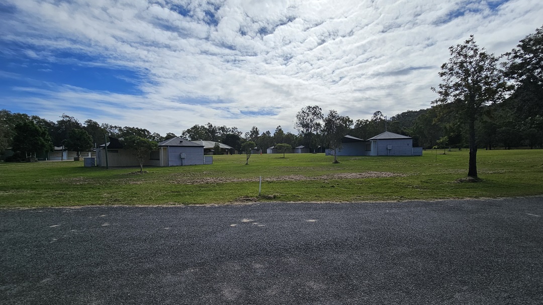 A grassy field with several small, light blue buildings scattered across it. Trees are visible in the background, surrounding the area. In the foreground, there is a gravel path or road. The sky is partly cloudy, casting a soft light over the scene. A grassy field with several small, light blue buildings scattered across it. Trees are visible in the background, surrounding the area. In the foreground, there is a gravel path or road. The sky is partly cloudy, casting a soft light over the scene.