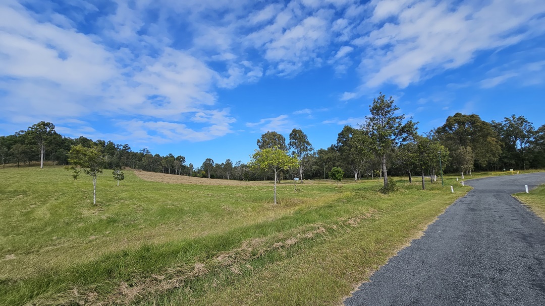 A landscape with a grassy field on the left, dotted with small trees. On the right, a gravel path curves through the scene, bordered by more trees. The background features a line of taller trees along the horizon. The sky is partly cloudy with patches of blue visible. A landscape with a grassy field on the left, dotted with small trees. On the right, a gravel path curves through the scene, bordered by more trees. The background features a line of taller trees along the horizon. The sky is partly cloudy with patches of blue visible.