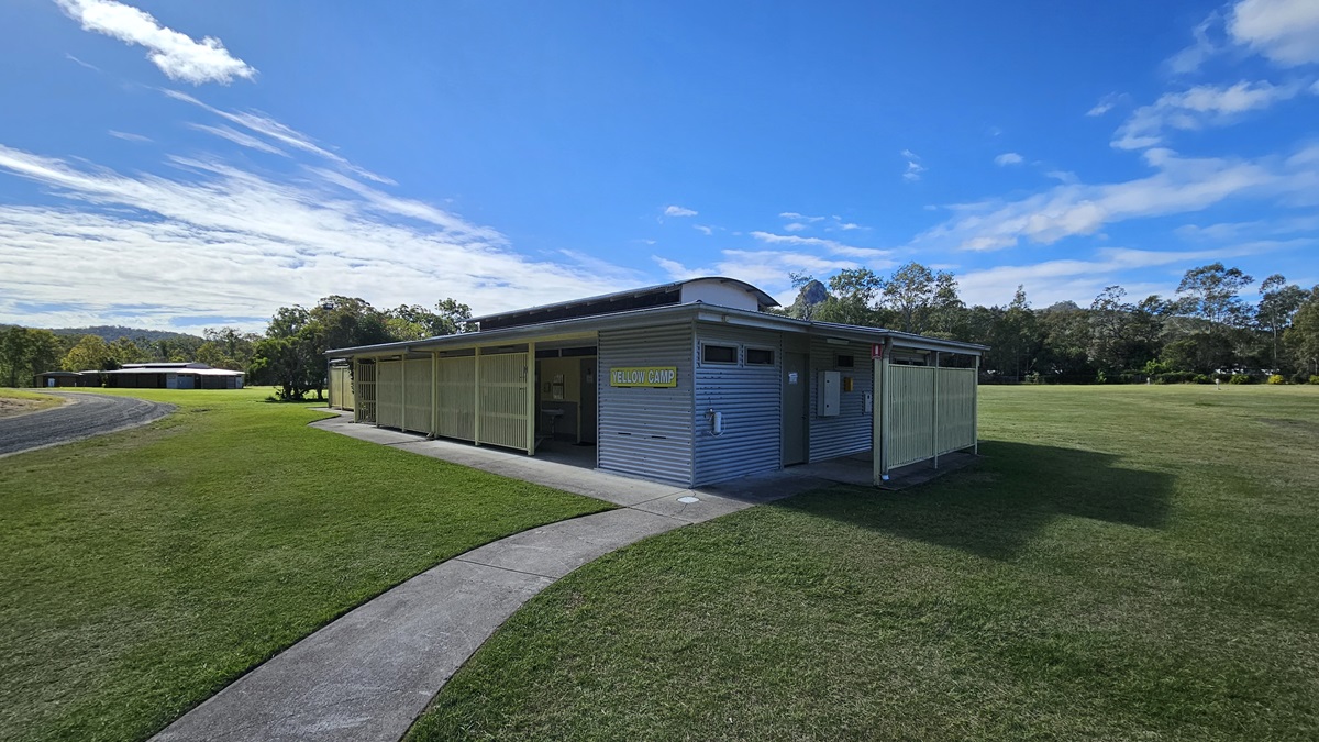 A single-storey building with a corrugated metal exterior is situated on a grassy field. The structure has a flat roof and a covered entrance. A concrete path leads to the building, curving from the foreground. In the background, there are trees and another similar building. The sky is clear with some clouds. A single-storey building with a corrugated metal exterior is situated on a grassy field. The structure has a flat roof and a covered entrance. A concrete path leads to the building, curving from the foreground. In the background, there are trees and another similar building. The sky is clear with some clouds.