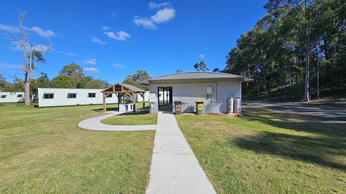 A grassy area with a paved path leading to a small building in the centre. The building has a corrugated metal exterior and is flanked by two large gas cylinders and bins. To the left, there is a covered picnic area with a wooden structure and a few portable cabins in the background. On the right, there are tall trees casting shadows on the grass. The sky is clear with a few clouds. A grassy area with a paved path leading to a small building in the centre. The building has a corrugated metal exterior and is flanked by two large gas cylinders and bins. To the left, there is a covered picnic area with a wooden structure and a few portable cabins in the background. On the right, there are tall trees casting shadows on the grass. The sky is clear with a few clouds.