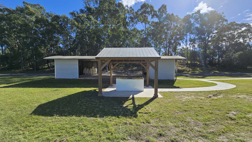 A small wooden shelter with a metal roof stands in the centre of a grassy area. In front of it is a white rectangular structure, possibly a table or bench. Behind the shelter, there is a larger building with a corrugated metal exterior and open sides, containing stacked chairs. A curved concrete path runs from the foreground to the right side of the image. Tall trees with dense foliage form the background. A small wooden shelter with a metal roof stands in the centre of a grassy area. In front of it is a white rectangular structure, possibly a table or bench. Behind the shelter, there is a larger building with a corrugated metal exterior and open sides, containing stacked chairs. A curved concrete path runs from the foreground to the right side of the image. Tall trees with dense foliage form the background.