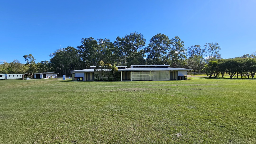A grassy field with a low, rectangular building in the centre, featuring a flat roof and large windows. The building is surrounded by trees in the background. To the left, there are smaller structures, possibly sheds or cabins. The sky is clear and blue. A grassy field with a low, rectangular building in the centre, featuring a flat roof and large windows. The building is surrounded by trees in the background. To the left, there are smaller structures, possibly sheds or cabins. The sky is clear and blue.