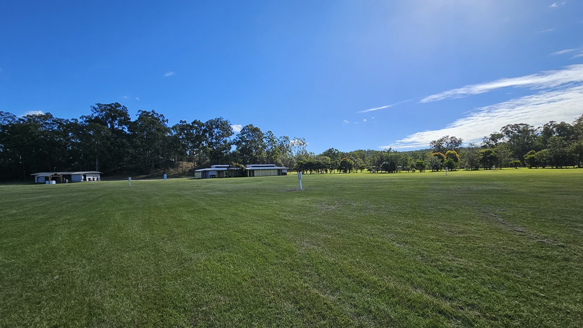 A wide, open grassy field stretches across the foreground, with a few small structures visible in the middle distance. In the background, a line of tall trees forms a dense border. The sky is clear with a few clouds, casting bright light over the scene. A wide, open grassy field stretches across the foreground, with a few small structures visible in the middle distance. In the background, a line of tall trees forms a dense border. The sky is clear with a few clouds, casting bright light over the scene.