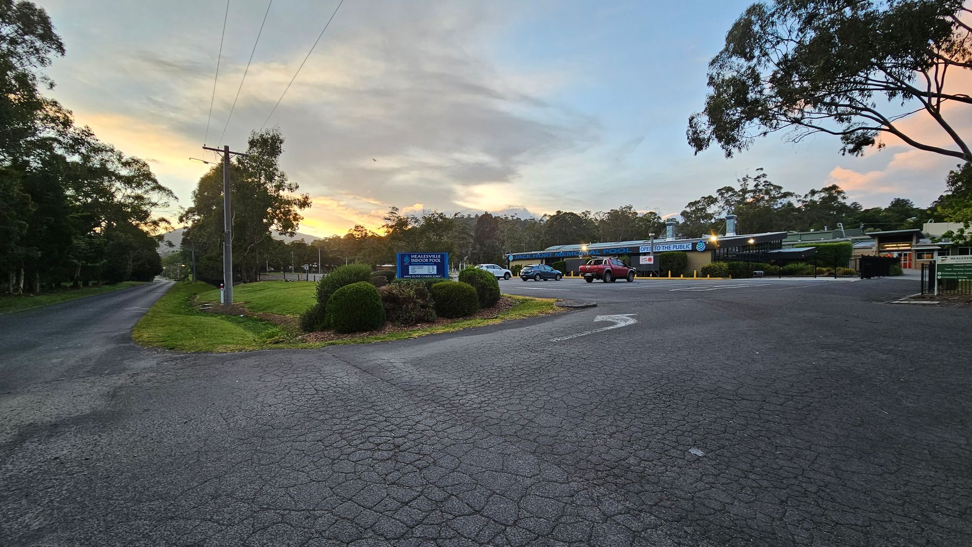 A paved road leads into the distance on the left, lined with trees and a power line. In the centre, a sign reads "Healesville Indoor Pool" with bushes around it. On the right, a parking area with a few cars is in front of a building labelled "Jack Hort Memorial Pool" with a sign indicating it's open to the public. More trees are visible in the background, and the sky has a mix of clouds and a hint of sunset colours. A paved road leads into the distance on the left, lined with trees and a power line. In the centre, a sign reads "Healesville Indoor Pool" with bushes around it. On the right, a parking area with a few cars is in front of a building labelled "Jack Hort Memorial Pool" with a sign indicating it's open to the public. More trees are visible in the background, and the sky has a mix of clouds and a hint of sunset colours.
