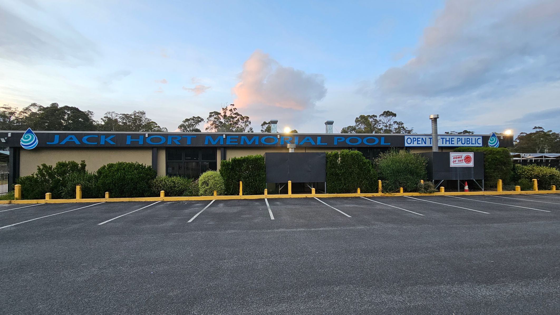 A building with a sign reading "Jack Hort Memorial Pool" in blue letters is centred in the image. The words "Open to the Public" are also visible. The foreground features an empty car park with marked spaces and yellow bollards. Bushes and greenery line the front of the building. In the background, there are trees and a partly cloudy sky. A building with a sign reading "Jack Hort Memorial Pool" in blue letters is centred in the image. The words "Open to the Public" are also visible. The foreground features an empty car park with marked spaces and yellow bollards. Bushes and greenery line the front of the building. In the background, there are trees and a partly cloudy sky.