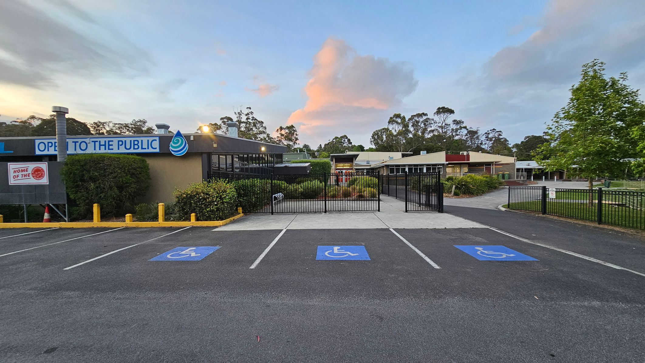 A parking area with three marked disabled parking spaces in the foreground. On the left, a building with a sign reading "OPEN TO THE PUBLIC" and a logo. The building is surrounded by bushes and yellow bollards. In the centre, a black metal gate leads to a pathway lined with greenery. On the right, another building and a tree are visible. The background features trees and a partly cloudy sky. A parking area with three marked disabled parking spaces in the foreground. On the left, a building with a sign reading "OPEN TO THE PUBLIC" and a logo. The building is surrounded by bushes and yellow bollards. In the centre, a black metal gate leads to a pathway lined with greenery. On the right, another building and a tree are visible. The background features trees and a partly cloudy sky.
