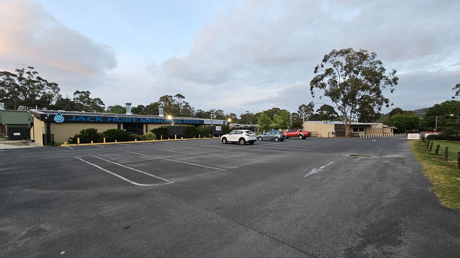 A large car park with a few vehicles, including a white SUV, a green car, and a red truck, is in the foreground. On the left, a building with a sign reading "Jack Hort Memorial Pool" is visible, surrounded by bushes. The background features tall trees and a cloudy sky. The ground is paved, and there is a grassy area with wooden posts on the right. A large car park with a few vehicles, including a white SUV, a green car, and a red truck, is in the foreground. On the left, a building with a sign reading "Jack Hort Memorial Pool" is visible, surrounded by bushes. The background features tall trees and a cloudy sky. The ground is paved, and there is a grassy area with wooden posts on the right.