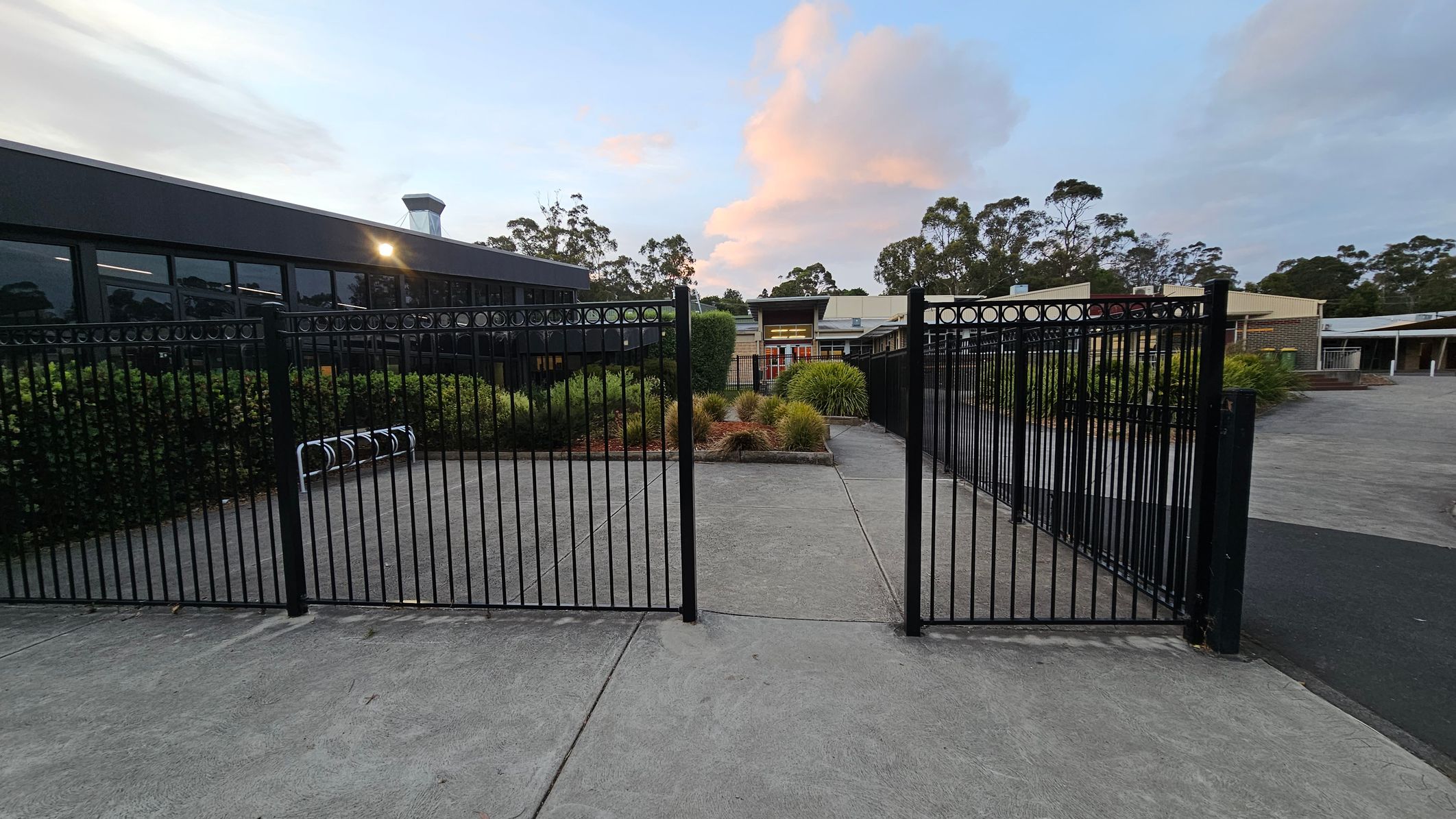 A black metal gate stands open on a concrete path. On the left, there's a building with large windows reflecting the surroundings, and a bike rack is visible. In the centre, there are landscaped bushes and plants. On the right, more plants line the path leading to another building in the background. Tall trees are visible in the distance. A black metal gate stands open on a concrete path. On the left, there's a building with large windows reflecting the surroundings, and a bike rack is visible. In the centre, there are landscaped bushes and plants. On the right, more plants line the path leading to another building in the background. Tall trees are visible in the distance.