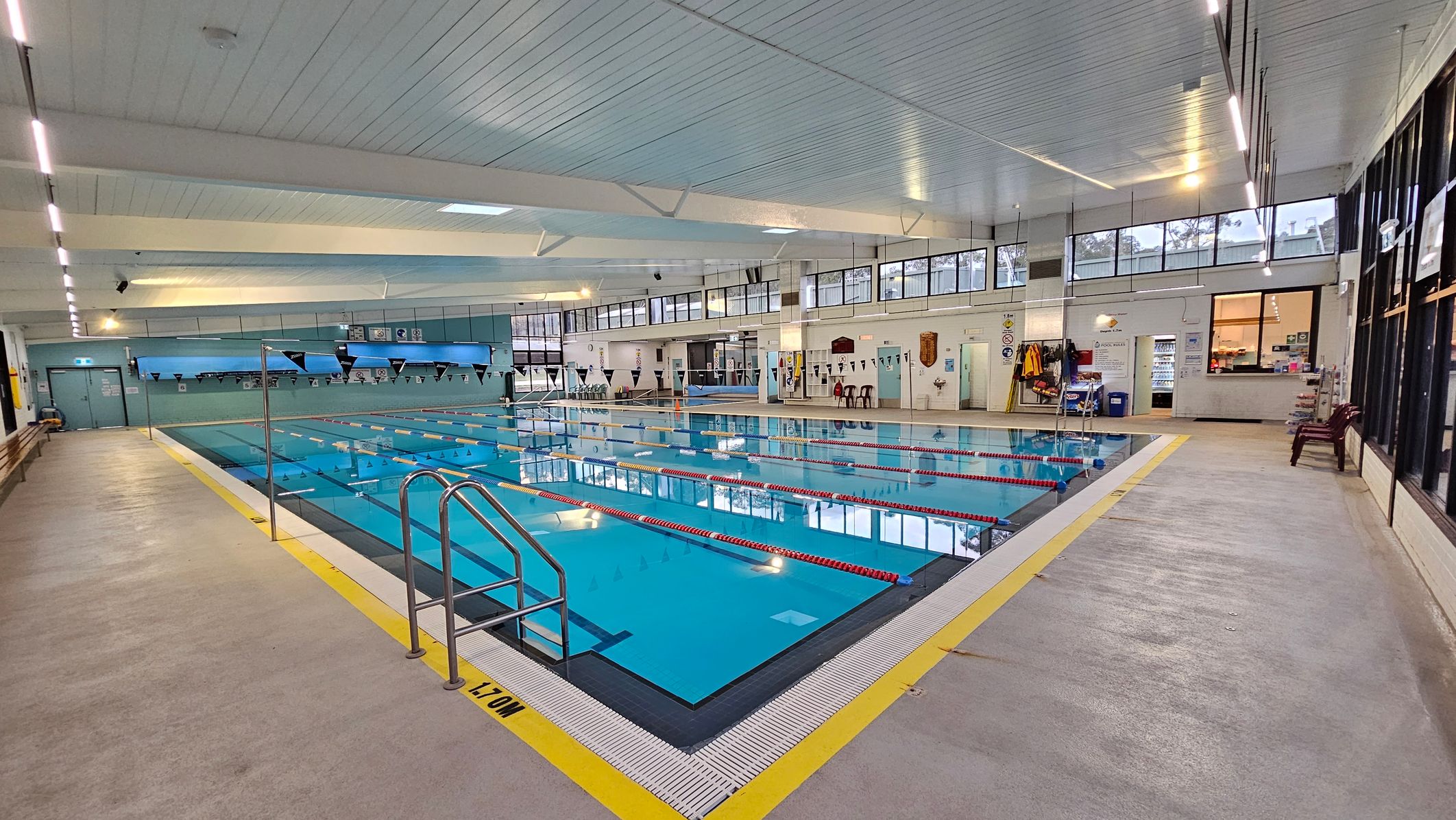 Indoor swimming pool with clear blue water and red lane dividers. On the left, a beige walkway with wooden benches runs alongside the pool. On the right, a similar walkway with maroon chairs and a vending machine. The back wall features large windows and a door. The ceiling is white with fluorescent lights, and the floor is a mix of beige and light purple. Indoor swimming pool with clear blue water and red lane dividers. On the left, a beige walkway with wooden benches runs alongside the pool. On the right, a similar walkway with maroon chairs and a vending machine. The back wall features large windows and a door. The ceiling is white with fluorescent lights, and the floor is a mix of beige and light purple.