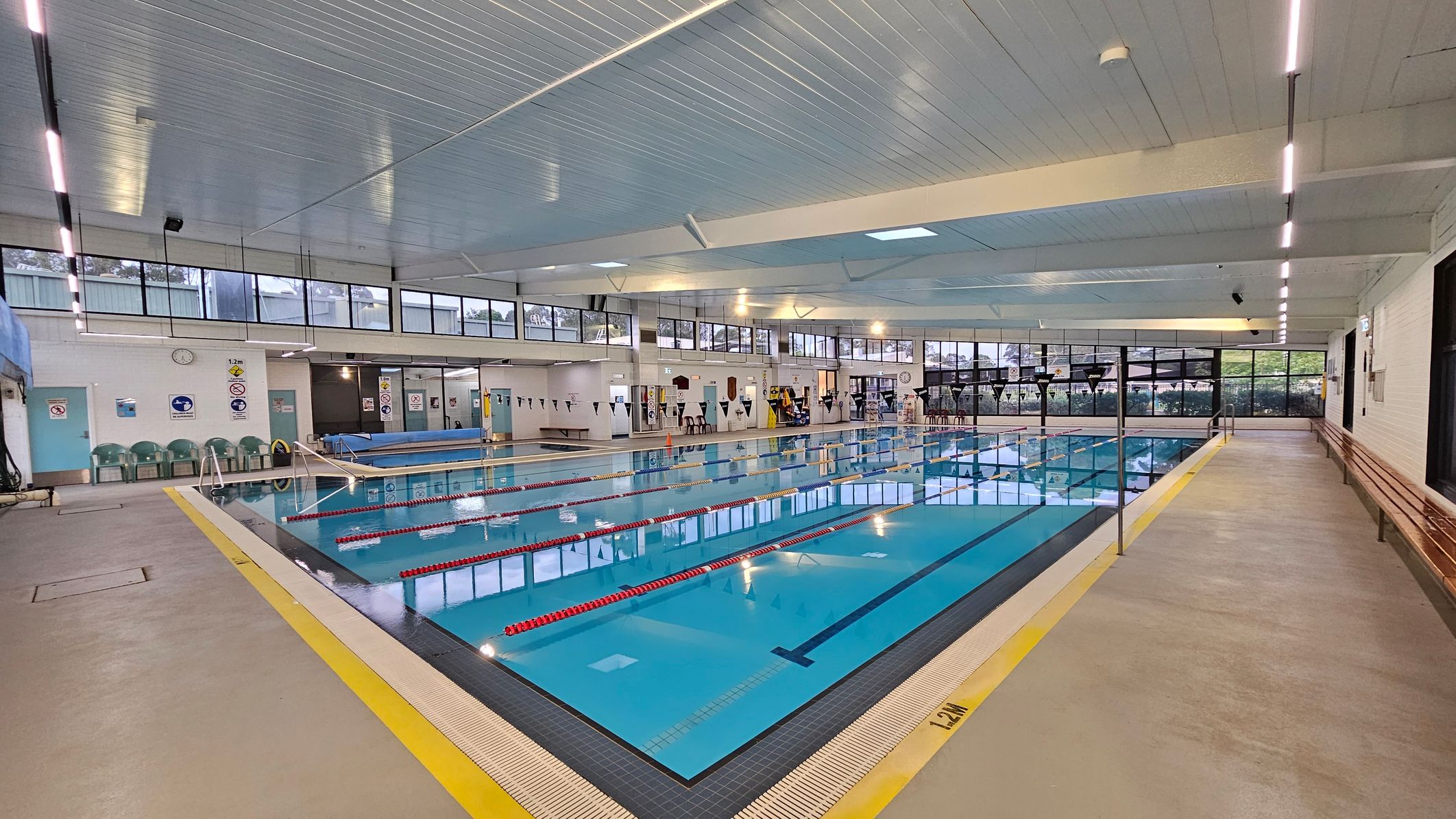 An indoor swimming pool with clear blue water and red lane dividers. On the left, there are green plastic chairs against a white wall with signs and a clock. The right side features a long wooden bench along the wall. The ceiling is white with strip lighting, and large windows line the upper walls, allowing natural light in. The floor is a light grey with a yellow safety line around the pool's edge. An indoor swimming pool with clear blue water and red lane dividers. On the left, there are green plastic chairs against a white wall with signs and a clock. The right side features a long wooden bench along the wall. The ceiling is white with strip lighting, and large windows line the upper walls, allowing natural light in. The floor is a light grey with a yellow safety line around the pool's edge.
