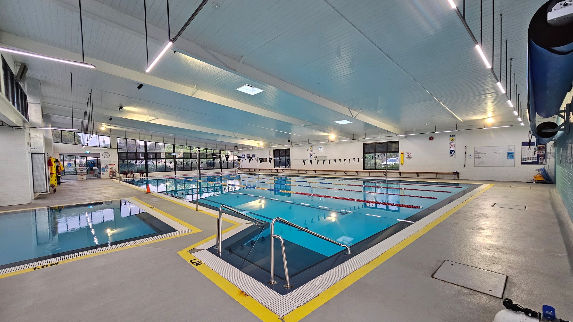 Indoor swimming pool area with a large main pool in the centre, marked with lane dividers. On the left, a smaller pool is visible. The floor is grey with yellow safety markings around the pool edges. The ceiling is light blue with long, narrow lights. Large windows line the left wall, allowing natural light in. On the right, pool equipment is stored on racks. A clock and safety signs are mounted on the far wall. Indoor swimming pool area with a large main pool in the centre, marked with lane dividers. On the left, a smaller pool is visible. The floor is grey with yellow safety markings around the pool edges. The ceiling is light blue with long, narrow lights. Large windows line the left wall, allowing natural light in. On the right, pool equipment is stored on racks. A clock and safety signs are mounted on the far wall.