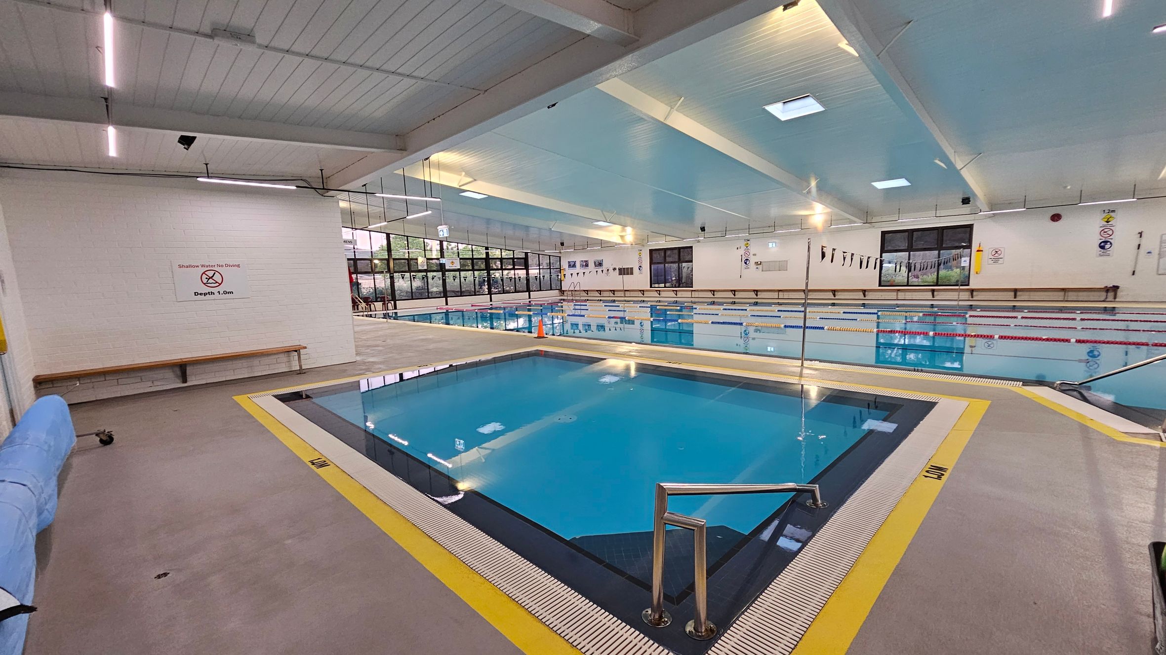 Indoor swimming pool area with a small, square pool in the foreground, surrounded by a yellow border and a metal handrail. A sign on the left wall reads "Shallow Water No Diving, Depth 1.0m." Behind it, a larger pool with lane dividers stretches across the room. The ceiling is high with exposed beams and skylights. Large windows line the far wall, allowing natural light to enter. A bench is positioned against the left wall. Indoor swimming pool area with a small, square pool in the foreground, surrounded by a yellow border and a metal handrail. A sign on the left wall reads "Shallow Water No Diving, Depth 1.0m." Behind it, a larger pool with lane dividers stretches across the room. The ceiling is high with exposed beams and skylights. Large windows line the far wall, allowing natural light to enter. A bench is positioned against the left wall.