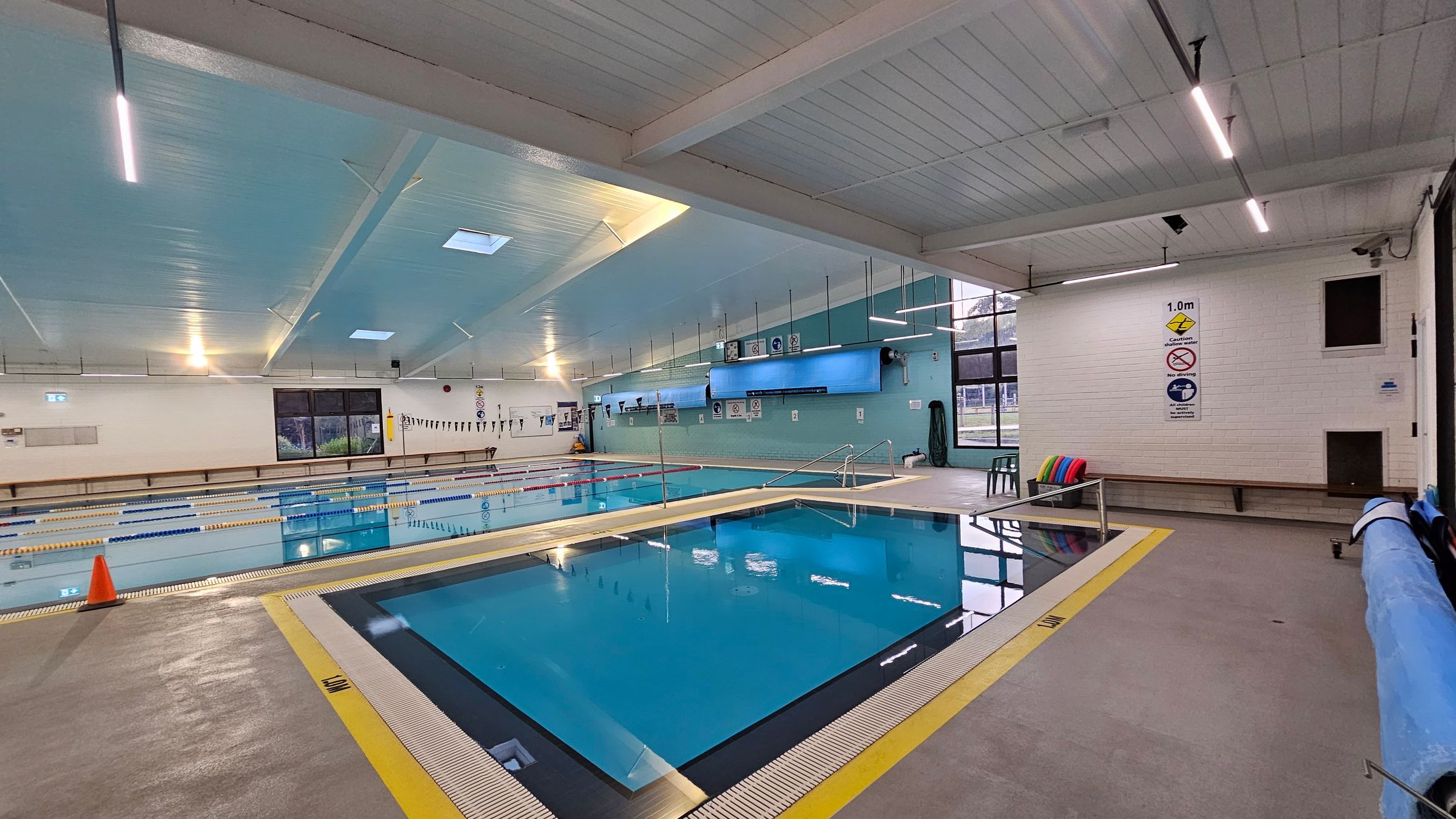 Indoor swimming pool area with two pools. The foreground pool is smaller, with a yellow safety line around it. The larger pool in the background has lane dividers and a cone on the deck. The walls are light blue and white, with safety signs and windows on the right. The ceiling is light blue with fluorescent lights. A bench with colourful floats is against the right wall, and a pool cover is rolled up on the right side. Indoor swimming pool area with two pools. The foreground pool is smaller, with a yellow safety line around it. The larger pool in the background has lane dividers and a cone on the deck. The walls are light blue and white, with safety signs and windows on the right. The ceiling is light blue with fluorescent lights. A bench with colourful floats is against the right wall, and a pool cover is rolled up on the right side.