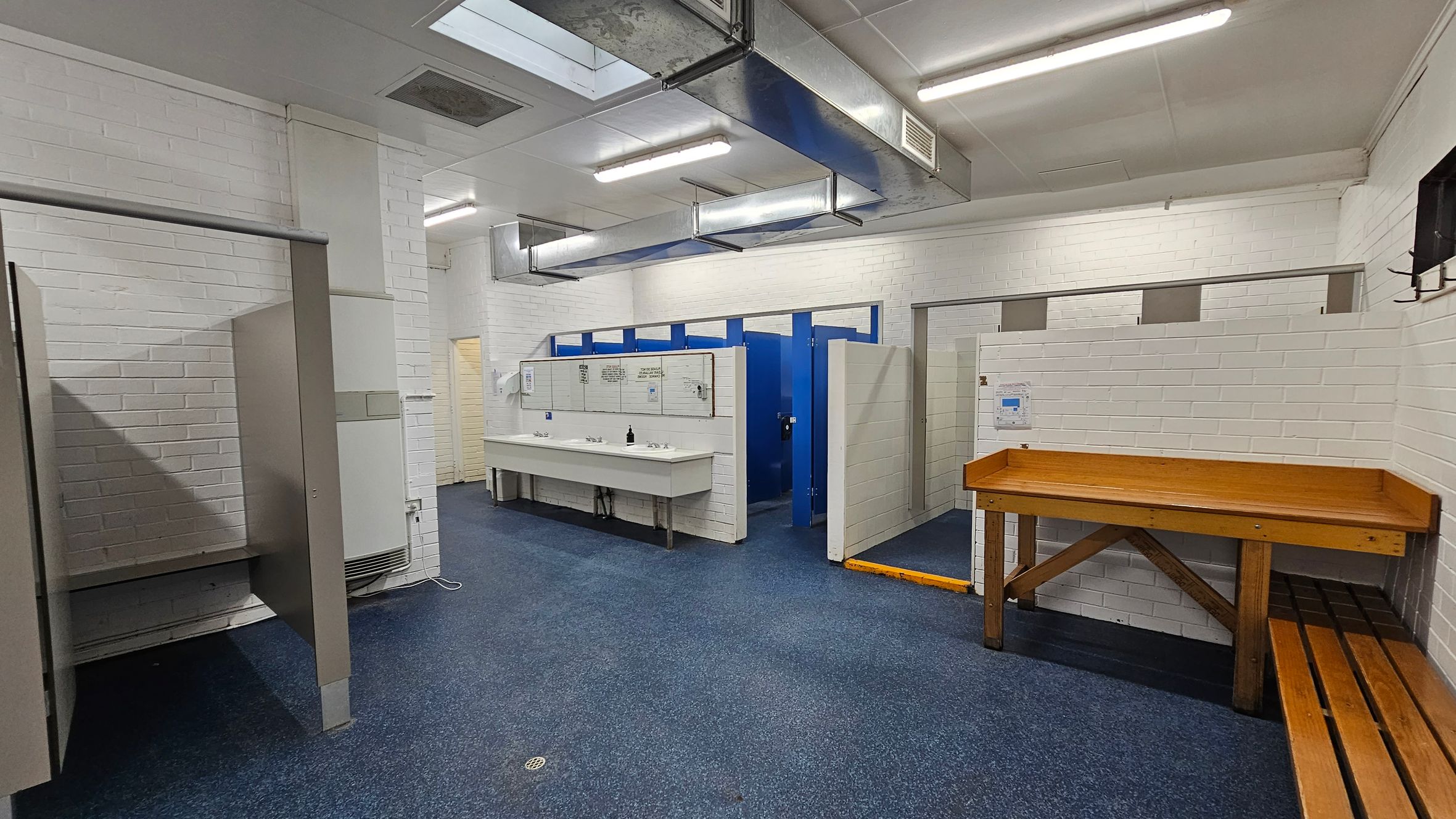 A changing room with white brick walls and a blue speckled floor. On the left, there are open cubicles with partitions. In the centre, a long white sink with multiple taps is mounted on the wall, above which are mirrors and notices. To the right, blue doors lead to toilet stalls. A wooden bench and table are positioned against the right wall. The ceiling has fluorescent lights and exposed ductwork. A changing room with white brick walls and a blue speckled floor. On the left, there are open cubicles with partitions. In the centre, a long white sink with multiple taps is mounted on the wall, above which are mirrors and notices. To the right, blue doors lead to toilet stalls. A wooden bench and table are positioned against the right wall. The ceiling has fluorescent lights and exposed ductwork.