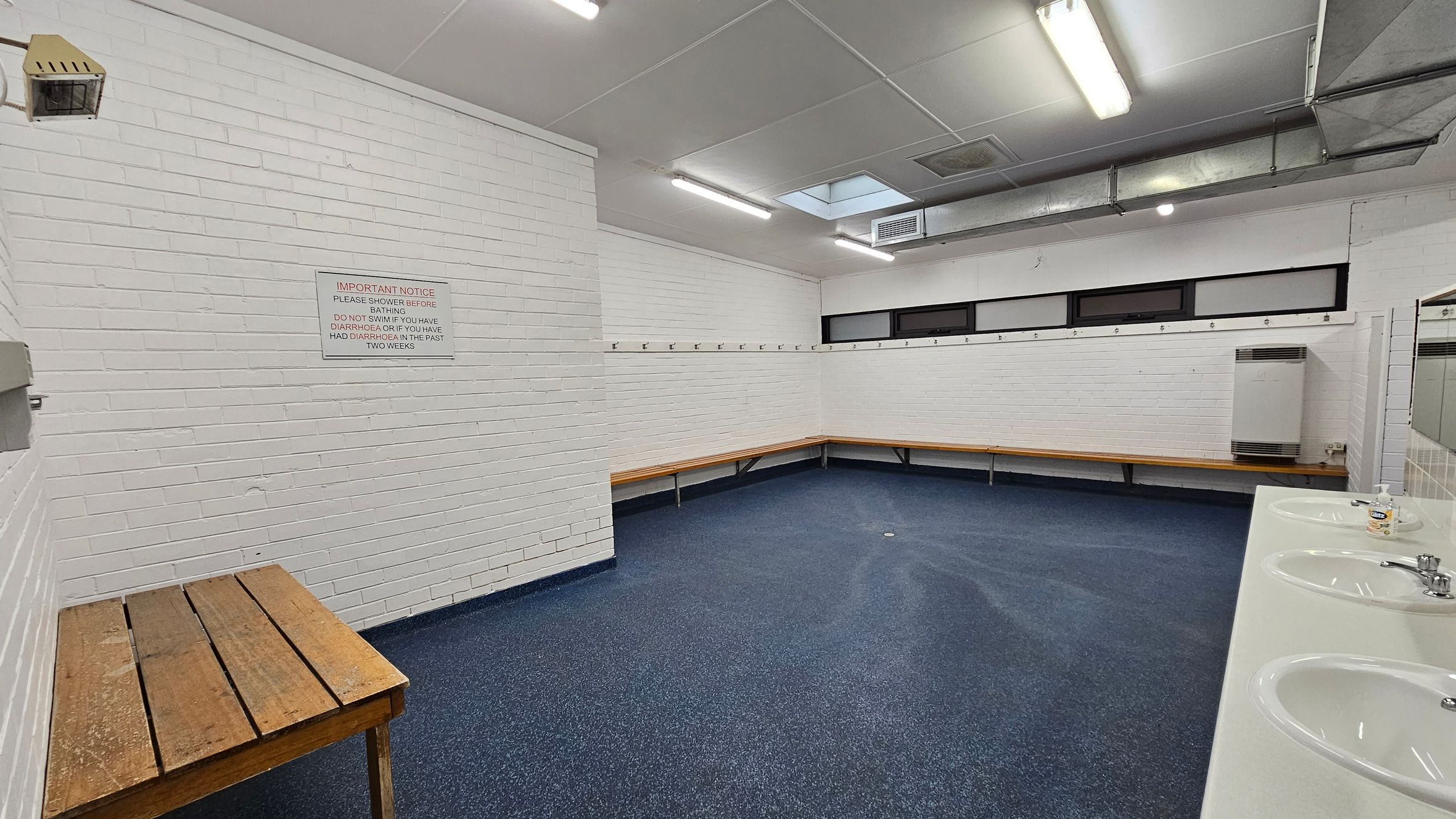 A changing room with white brick walls and a blue speckled floor. On the left, there's a wooden bench and a wall-mounted notice. The right side features a row of sinks with mirrors above them. Along the back wall, there are wooden benches and hooks for hanging items. The ceiling has fluorescent lights and a ventilation duct. A changing room with white brick walls and a blue speckled floor. On the left, there's a wooden bench and a wall-mounted notice. The right side features a row of sinks with mirrors above them. Along the back wall, there are wooden benches and hooks for hanging items. The ceiling has fluorescent lights and a ventilation duct.