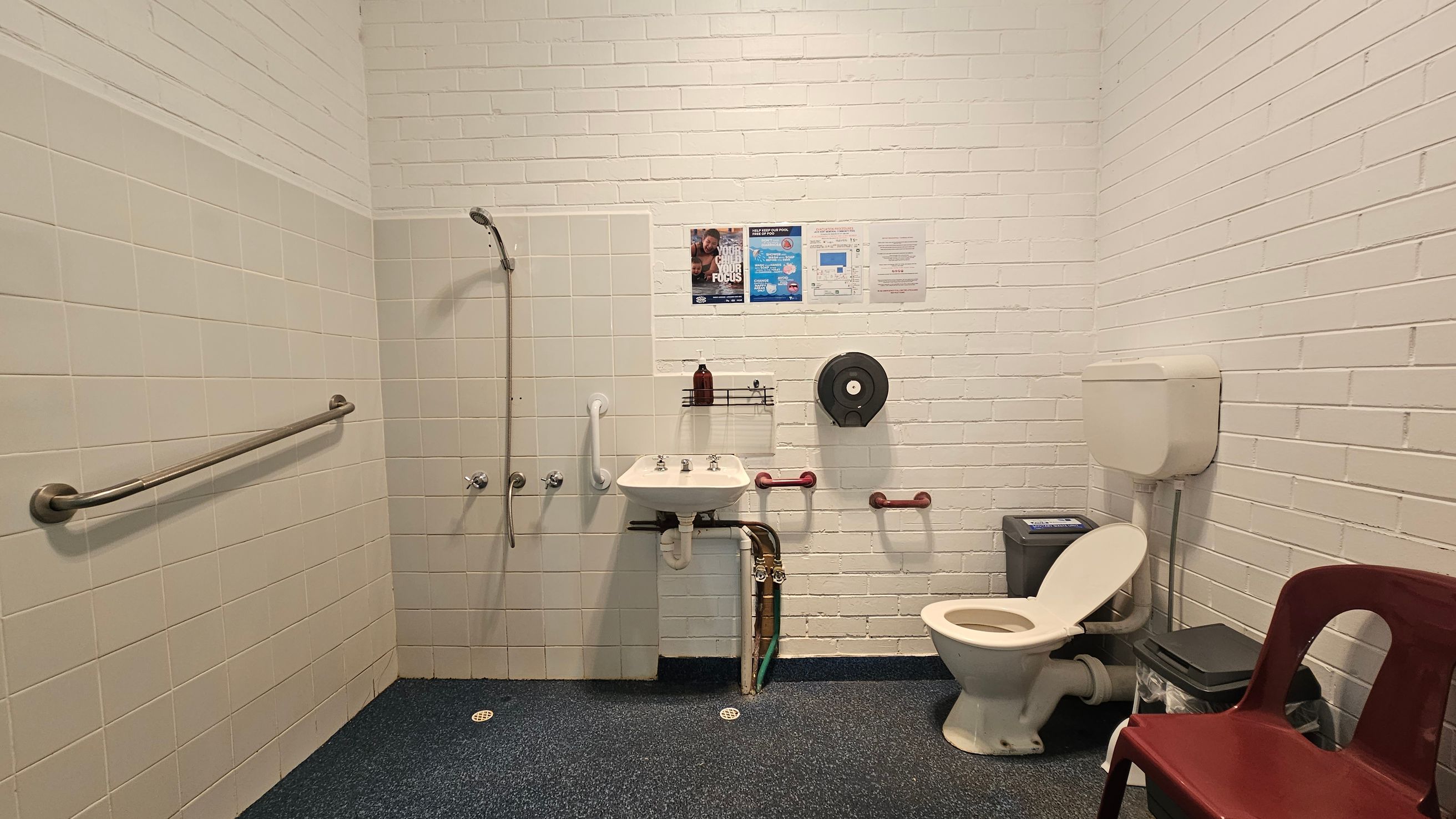 A bathroom with white brick walls and a dark speckled floor. On the left, there's a shower area with a handheld showerhead and a horizontal grab bar. In the centre, a small sink with a soap dispenser above it is mounted on the wall, with a toilet paper dispenser nearby. On the right, a toilet with a cistern is next to a green bin and a red plastic chair. Several posters and notices are on the wall above the sink. A bathroom with white brick walls and a dark speckled floor. On the left, there's a shower area with a handheld showerhead and a horizontal grab bar. In the centre, a small sink with a soap dispenser above it is mounted on the wall, with a toilet paper dispenser nearby. On the right, a toilet with a cistern is next to a green bin and a red plastic chair. Several posters and notices are on the wall above the sink.