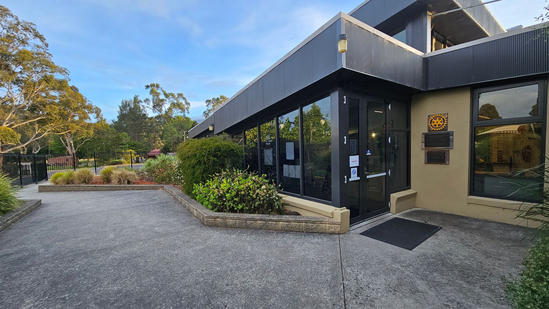 A modern building with large glass windows and a dark exterior is on the right. A plaque with a rotary emblem is mounted on the wall near the entrance. The entrance has a black door with a small ramp leading up to it. On the left, there is a paved pathway bordered by low brick walls and lush greenery, including bushes and small trees. In the background, a black metal fence and more trees are visible, creating a serene, landscaped setting. A modern building with large glass windows and a dark exterior is on the right. A plaque with a rotary emblem is mounted on the wall near the entrance. The entrance has a black door with a small ramp leading up to it. On the left, there is a paved pathway bordered by low brick walls and lush greenery, including bushes and small trees. In the background, a black metal fence and more trees are visible, creating a serene, landscaped setting.