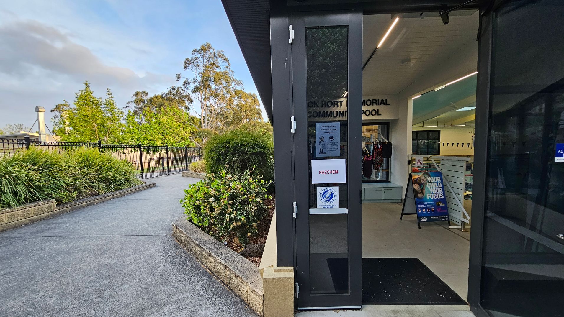 On the left, there's a pathway bordered by lush green bushes and a black metal fence. In the background, tall trees are visible. On the right, a partially open door leads into a building with signs, including one that says "HAZCHEM." Inside, there's a display of swimwear and a poster with the text "YOUR CHILD YOUR FOCUS." The ceiling has recessed lighting, and the floor is light-coloured. On the left, there's a pathway bordered by lush green bushes and a black metal fence. In the background, tall trees are visible. On the right, a partially open door leads into a building with signs, including one that says "HAZCHEM." Inside, there's a display of swimwear and a poster with the text "YOUR CHILD YOUR FOCUS." The ceiling has recessed lighting, and the floor is light-coloured.