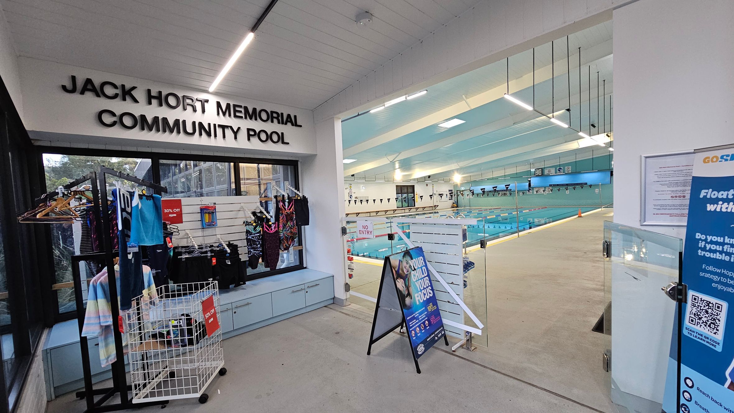 On the left, there is a display of swimwear on hangers and a basket with a "30% Off" sign. Above, the sign reads "Jack Hort Memorial Community Pool." On the right, a glass door leads to an indoor swimming pool with lanes and flags. A stand with a poster and a blue sign with safety information is visible. The ceiling has linear lights, and the floor is light-coloured. On the left, there is a display of swimwear on hangers and a basket with a "30% Off" sign. Above, the sign reads "Jack Hort Memorial Community Pool." On the right, a glass door leads to an indoor swimming pool with lanes and flags. A stand with a poster and a blue sign with safety information is visible. The ceiling has linear lights, and the floor is light-coloured.
