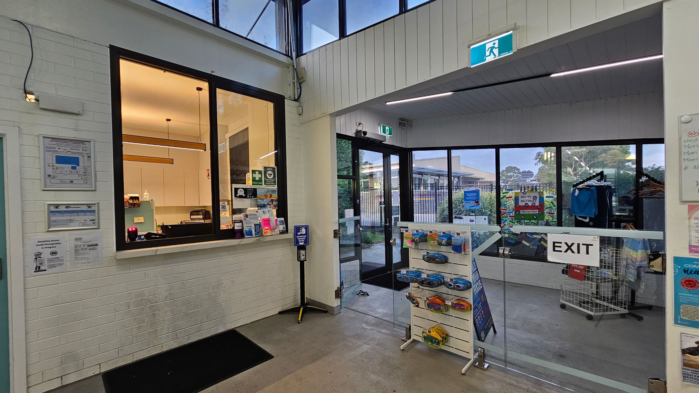 A reception area with a service window on the left, featuring a counter with various items and a lit interior. Notices and signs are displayed on the wall. In the centre, a hand sanitiser dispenser is mounted on a stand. To the right, a glass exit door leads outside, with a rack of clothing and a display stand with packaged items nearby. The ceiling has recessed lighting, and the floor is tiled. An emergency exit sign is visible above the door. A reception area with a service window on the left, featuring a counter with various items and a lit interior. Notices and signs are displayed on the wall. In the centre, a hand sanitiser dispenser is mounted on a stand. To the right, a glass exit door leads outside, with a rack of clothing and a display stand with packaged items nearby. The ceiling has recessed lighting, and the floor is tiled. An emergency exit sign is visible above the door.
