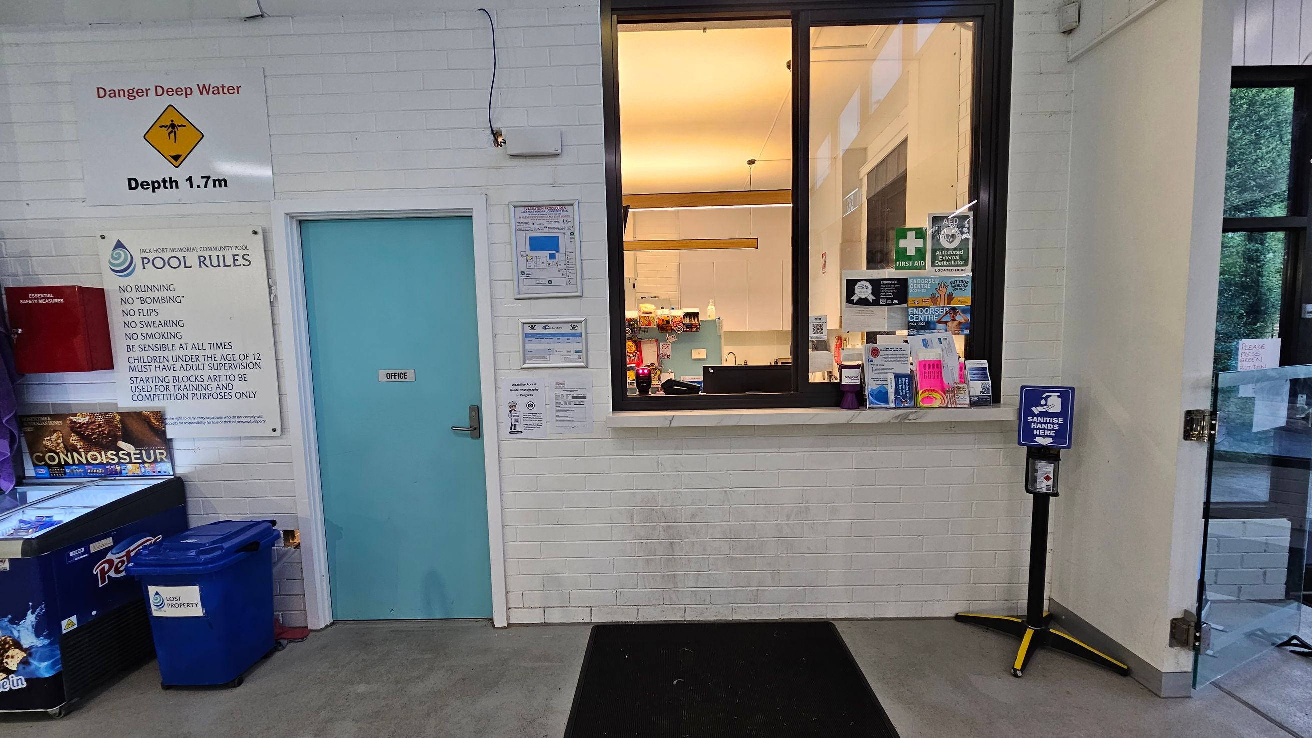 A white brick wall with a blue door labelled "Office" on the left. Above the door, a sign warns of deep water with a depth of 1.7m. Next to it, a board lists pool rules. Below, there's a freezer with "Connoisseur" branding and a blue bin marked "Lost Property". On the right, a window shows an office interior with various notices and a first aid sign. A hand sanitiser station is positioned near a glass door on the far right. The floor is grey with a black mat. A white brick wall with a blue door labelled "Office" on the left. Above the door, a sign warns of deep water with a depth of 1.7m. Next to it, a board lists pool rules. Below, there's a freezer with "Connoisseur" branding and a blue bin marked "Lost Property". On the right, a window shows an office interior with various notices and a first aid sign. A hand sanitiser station is positioned near a glass door on the far right. The floor is grey with a black mat.