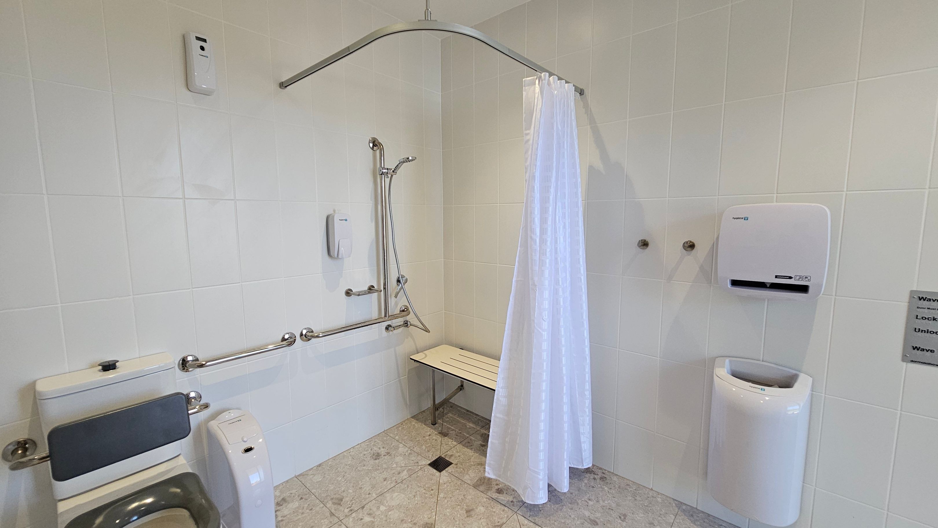 A bathroom with white tiled walls and a light-coloured tiled floor. On the left, there is a toilet with a backrest and grab bars. In the centre, a shower area features a handheld showerhead, grab bars, and a foldable bench. A white shower curtain hangs from a curved rod. On the right, a wall-mounted hand dryer and a paper towel dispenser are visible. A small sign with instructions is also present on the right wall.