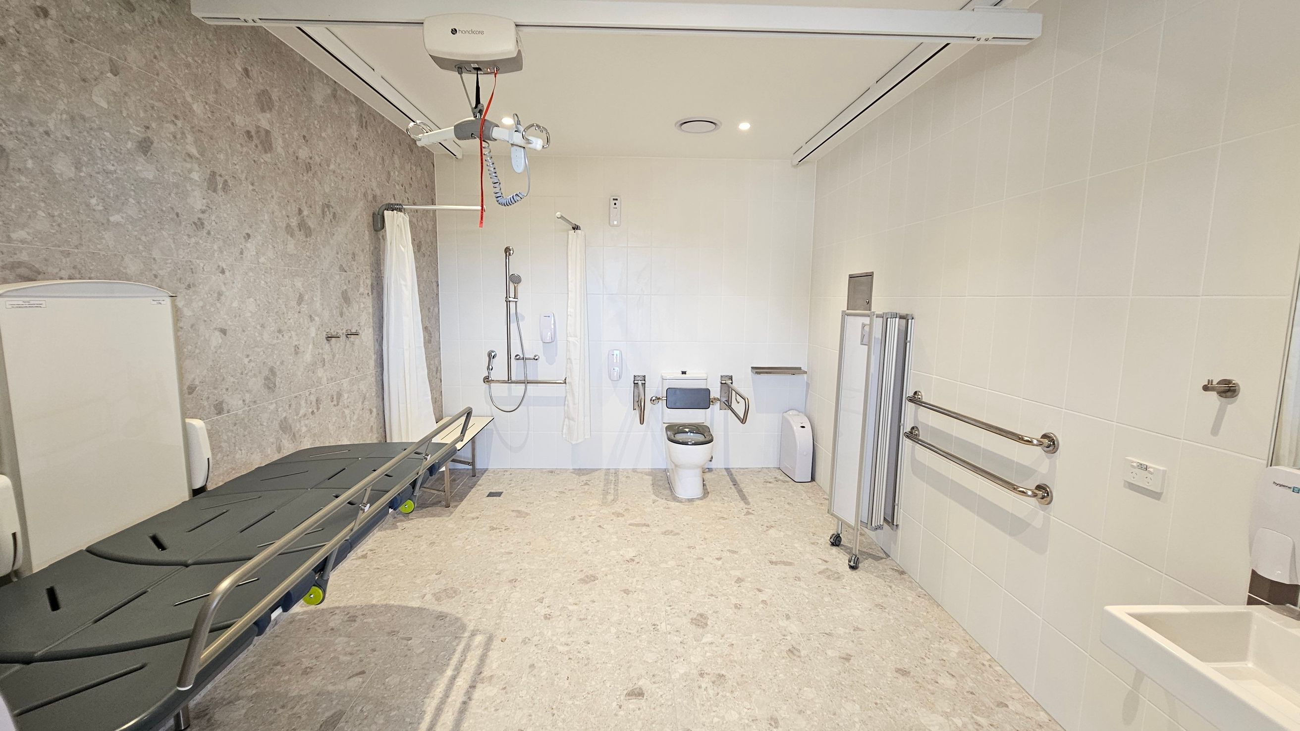 A spacious, accessible bathroom with a beige tiled floor and light-coloured walls. On the left, a large, adjustable changing table with side rails is positioned against the wall. In the centre, a toilet with support bars on either side is visible, along with a shower area equipped with a curtain and handrails. On the right, there are additional support bars and a small sink. Ceiling-mounted equipment for assistance is present, and a privacy screen is folded nearby.