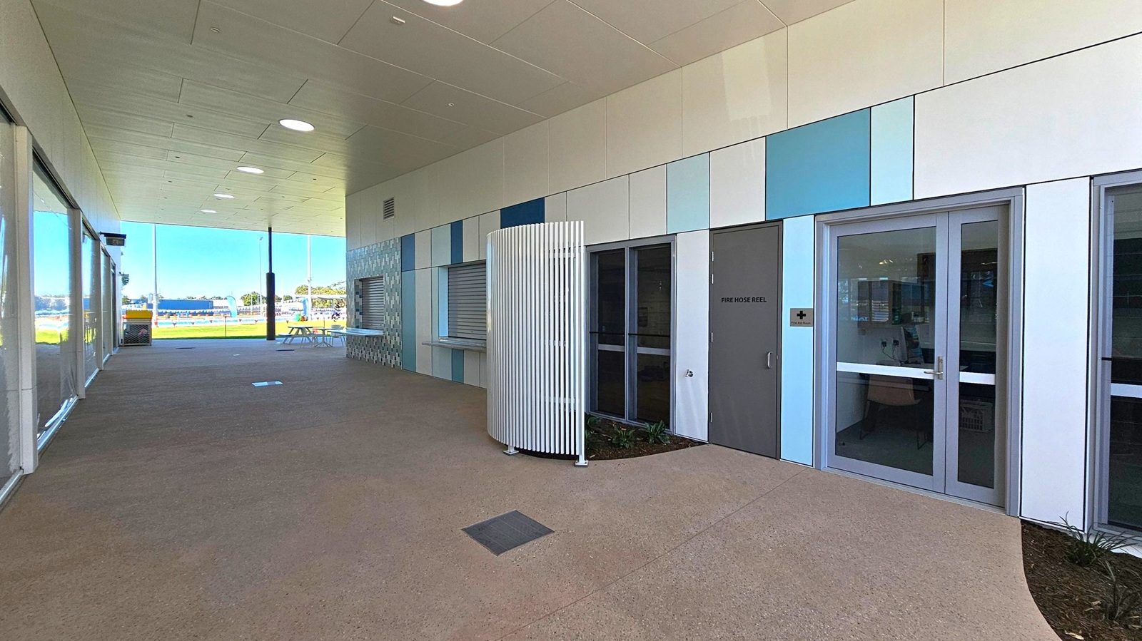 A covered outdoor area with a tiled ceiling featuring circular lights. The right side has a wall with a mix of blue and grey panels, a door marked "Fire Hose Reel," and a glass door with a medical cross sign. A curved white slatted structure is in front of the wall. The left side has large glass windows, and the floor is a mix of concrete and textured surfaces. In the background, there is a view of a grassy area with a picnic table and a distant structure.