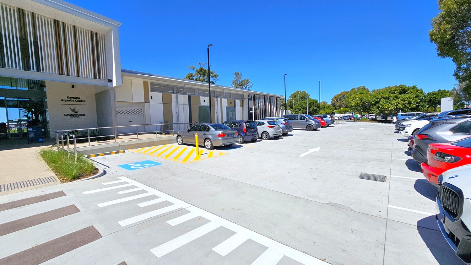 On the left, a modern building with vertical slats and signage for Kawana Aquatic Centre. In the middle, a row of parked cars, including a silver and a red car, with a marked disabled parking space in front. On the right, more parked cars and a line of trees in the background. The ground is a light grey concrete with white and yellow markings.