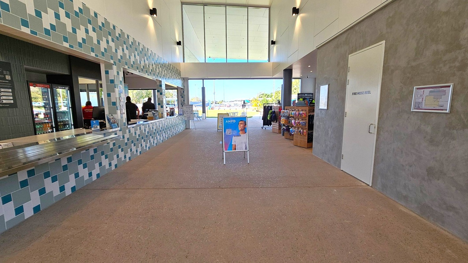 A spacious indoor area with a tiled counter on the left, featuring a mosaic pattern in shades of blue and grey. Behind the counter, there are windows with people visible inside. On the right, a grey and green textured wall with a door labelled "Fire Hose Reel" and a wall-mounted map. In the centre, a sign with a person’s image stands on a speckled floor. The background shows large glass windows and doors leading outside, with a view of trees and a clear sky.