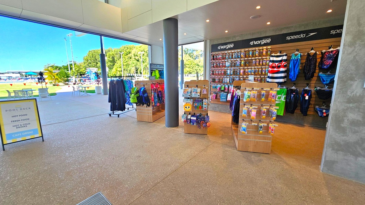 Inside a shop with a polished stone floor, there are several wooden display stands in the centre showcasing swimwear and accessories. On the right, a wall features hanging swim gear and branded items. A large window on the left reveals an outdoor area with tables, chairs, and a view of a swimming pool surrounded by greenery. A sign near the entrance advertises food and drinks. The ceiling is modern with recessed lighting.