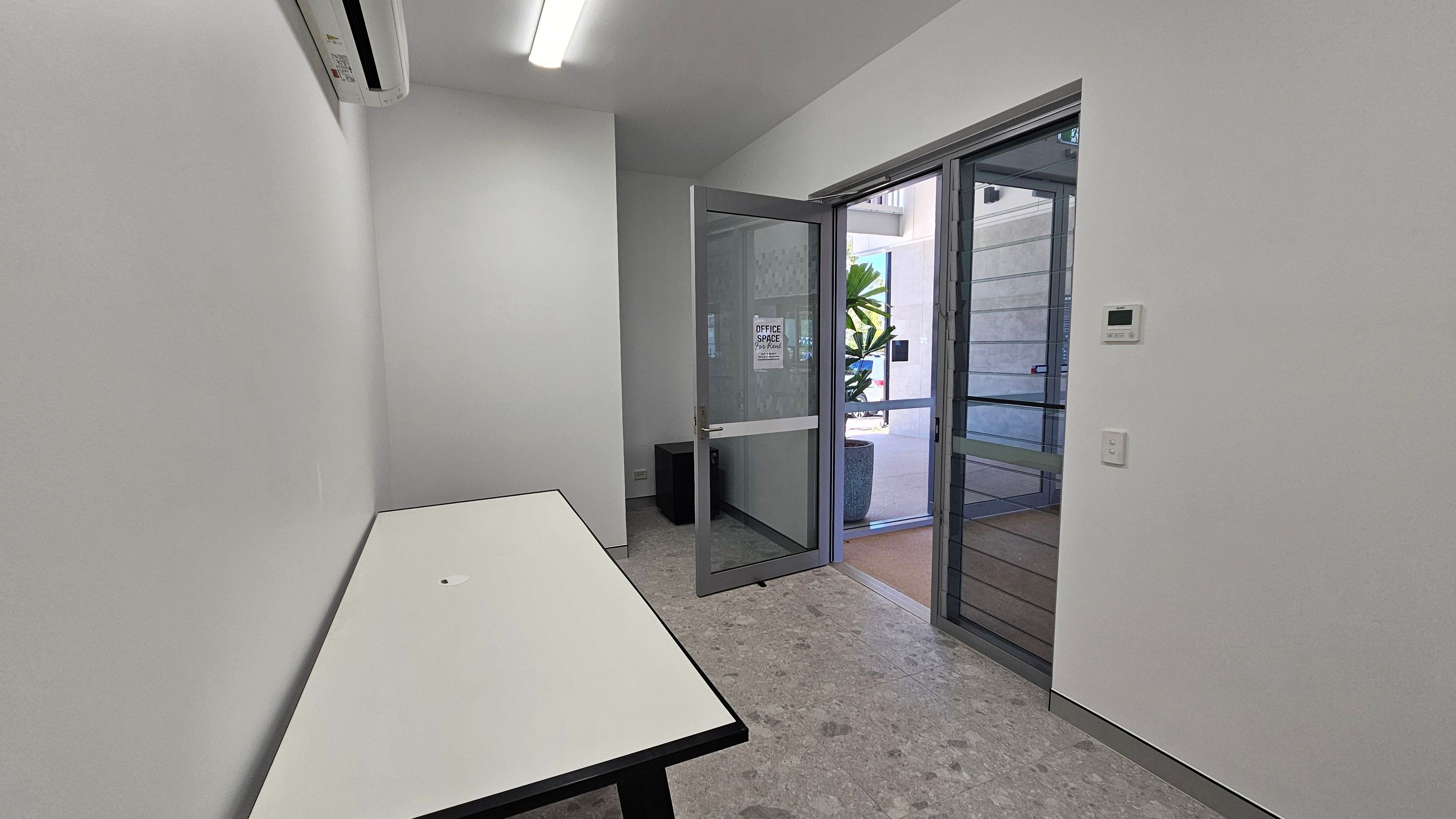 A small, narrow room with a white table on the left side. The floor is grey with a speckled pattern. On the right, a glass door is open, leading outside where a large potted plant is visible. Above the door, a fluorescent light fixture is mounted on the ceiling. An air conditioning unit is installed high on the left wall. A thermostat and a light switch are on the right wall. The walls are plain and white.