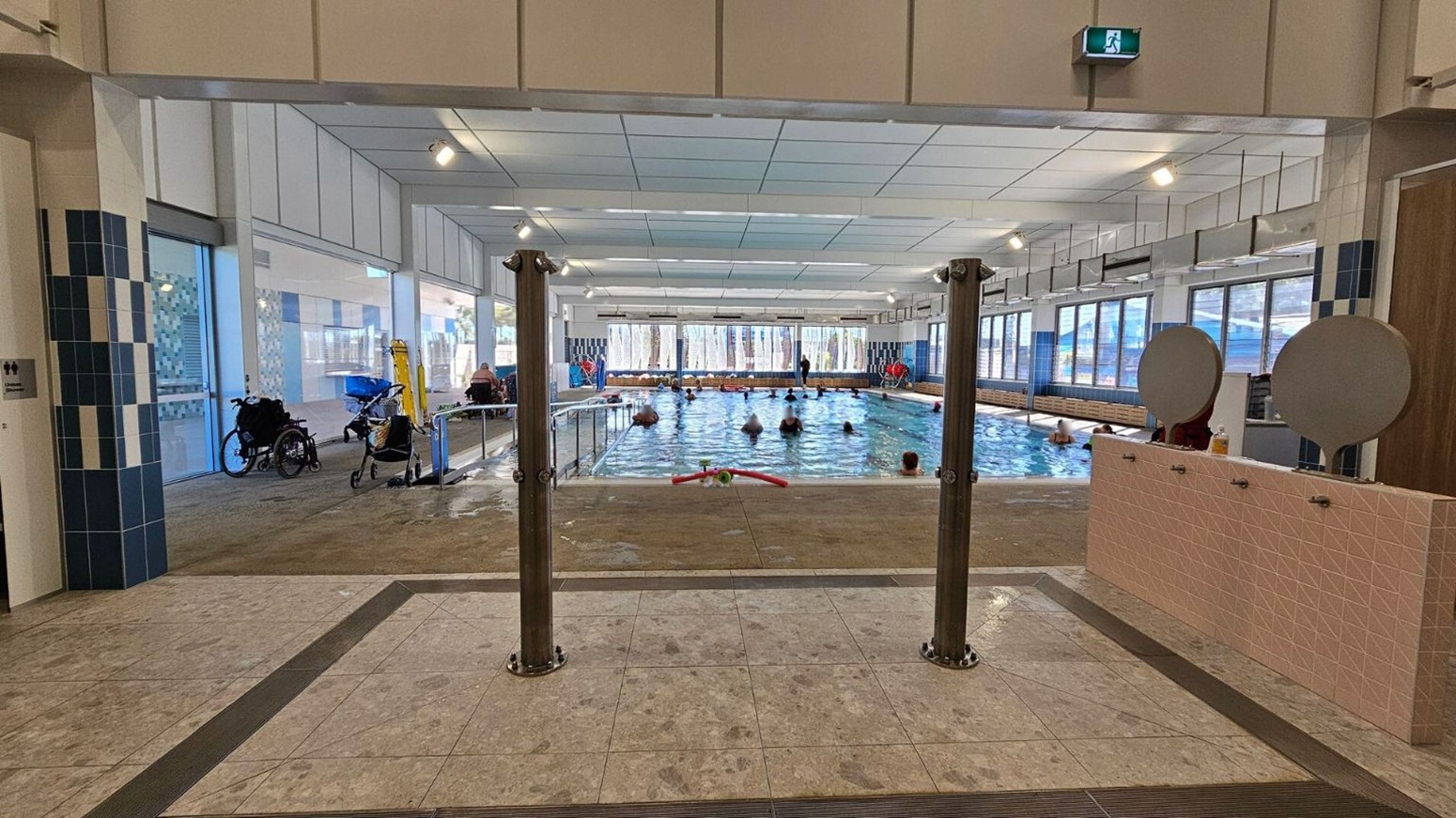 Indoor swimming pool with several people in the water. The pool is surrounded by large windows on the left and right, allowing natural light to enter. In the foreground, there are two wooden posts and a tiled floor with wet footprints. On the left, a wheelchair and a stroller are visible near the windows. On the right, there are two circular mirrors mounted on a tiled wall. The ceiling is white with recessed lighting, and an exit sign is visible above.
