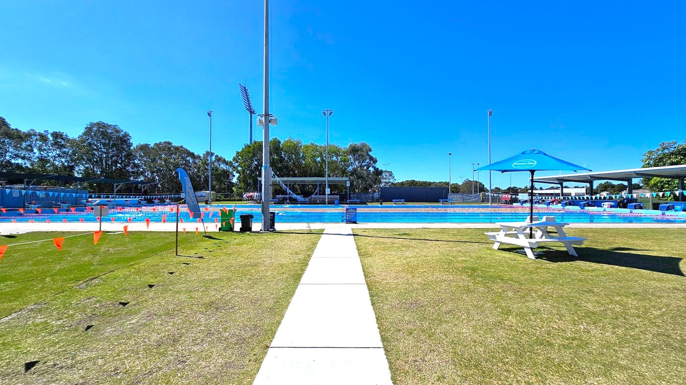 A swimming pool area with a concrete path leading to the pool in the centre. On the left, there are orange triangular flags and a blue flag on a pole. On the right, a white picnic table with a blue umbrella is on the grass. The pool is surrounded by blue and white bunting. In the background, there are trees and tall floodlights. The ground is a mix of grass and concrete.