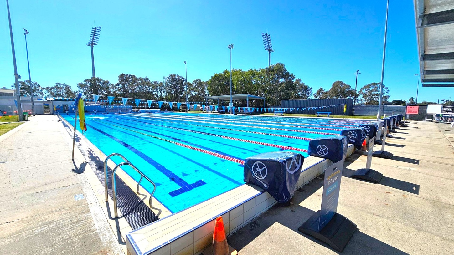 An outdoor swimming pool with multiple lanes, each marked by red and white lane dividers. Starting blocks are positioned at the pool's edge, covered with blue padding. A metal ladder is on the left side. The pool deck is concrete, with a traffic cone nearby. Tall floodlights and trees are visible in the background. A shaded seating area is on the far side, and a sign on the right reads "Lap Swimming in Progress."
