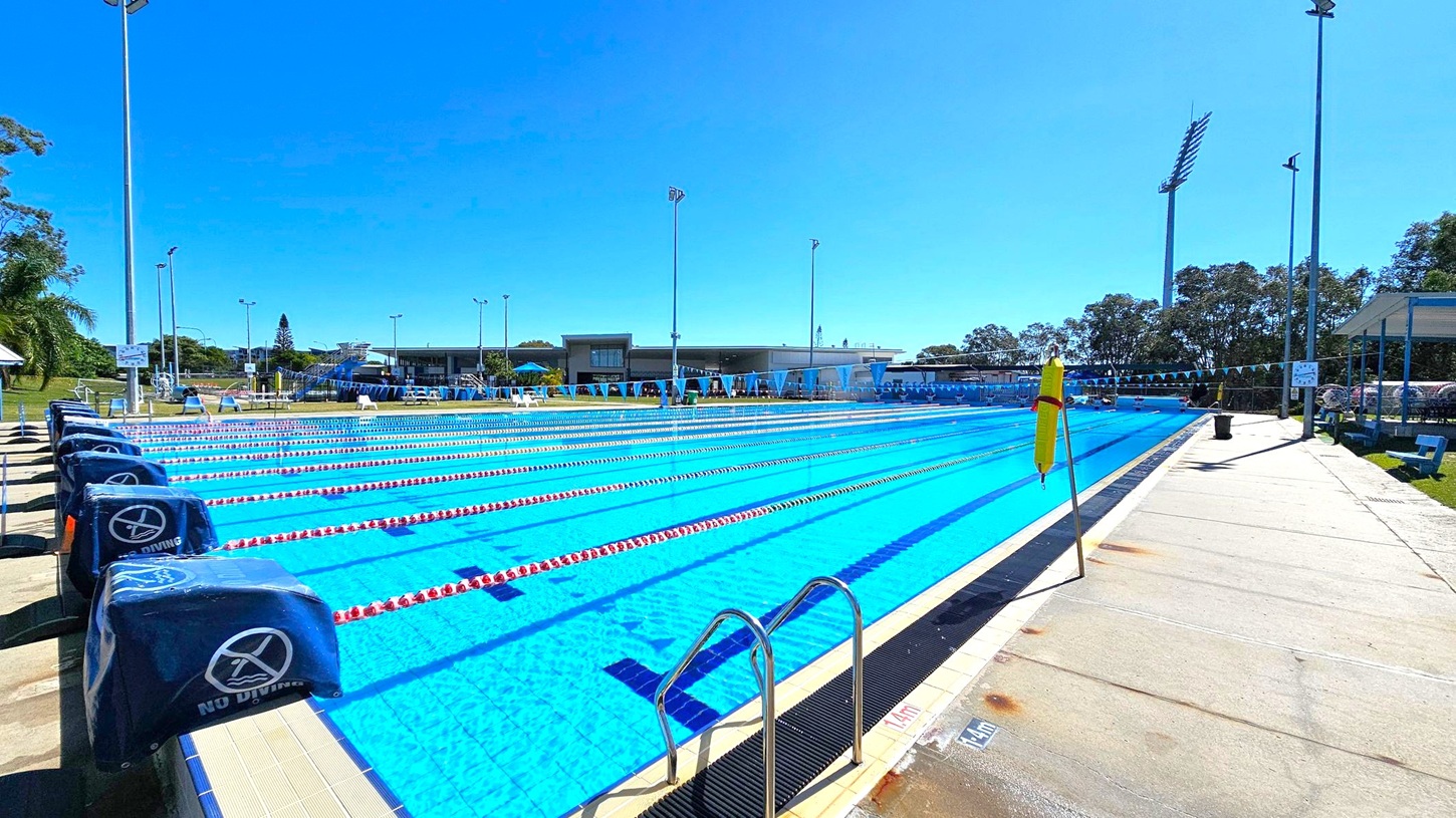 An outdoor swimming pool with multiple lanes, separated by red and white lane dividers. On the left, there are starting blocks covered with blue padding marked "No Diving." The poolside area is concrete, with a metal ladder on the right. In the background, there are trees, a building with a flat roof, and several tall floodlights. A yellow flag is positioned near the pool's edge. The area is well-lit, suggesting a sunny day.